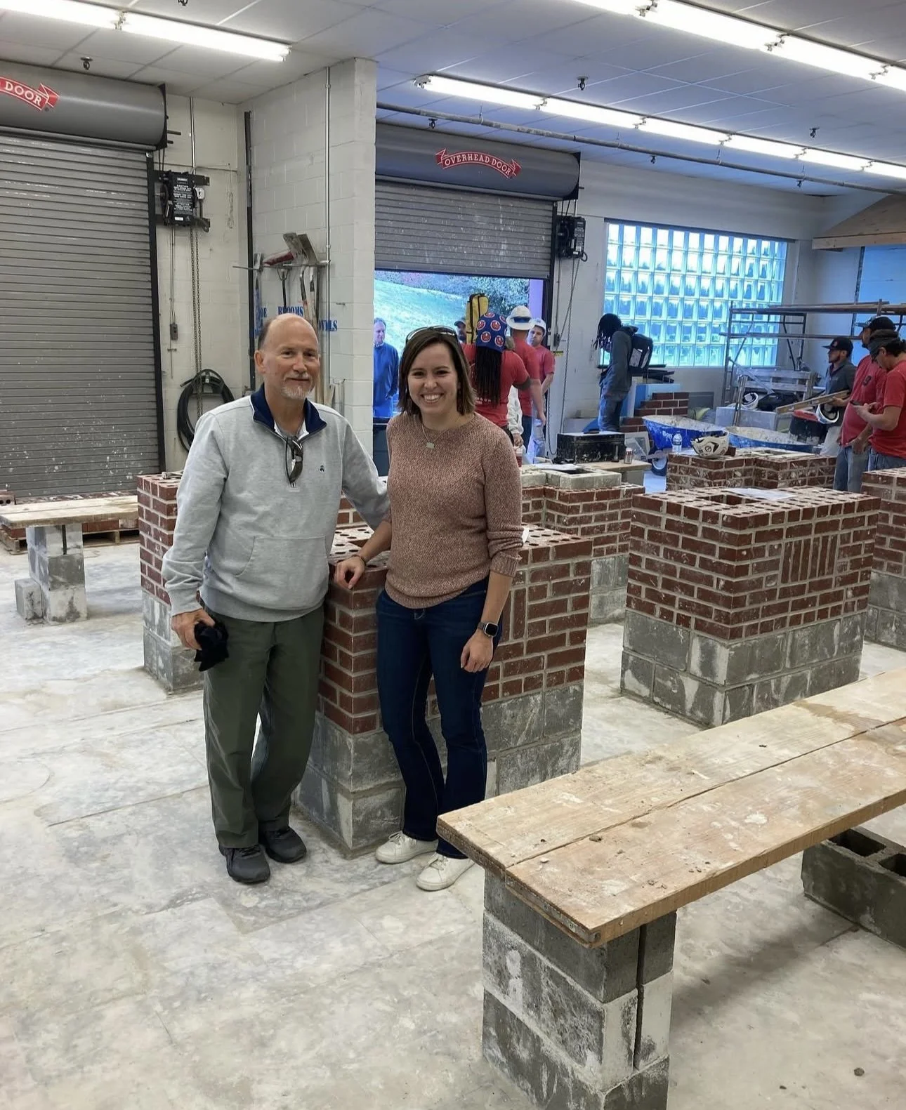 Two people standing inside a workshop, posing for a photo. The man on the left has a beard and is wearing a light gray sweatshirt and green pants. The woman on the right has short brown hair, is smiling, and wearing a brown sweater and dark jeans. Th