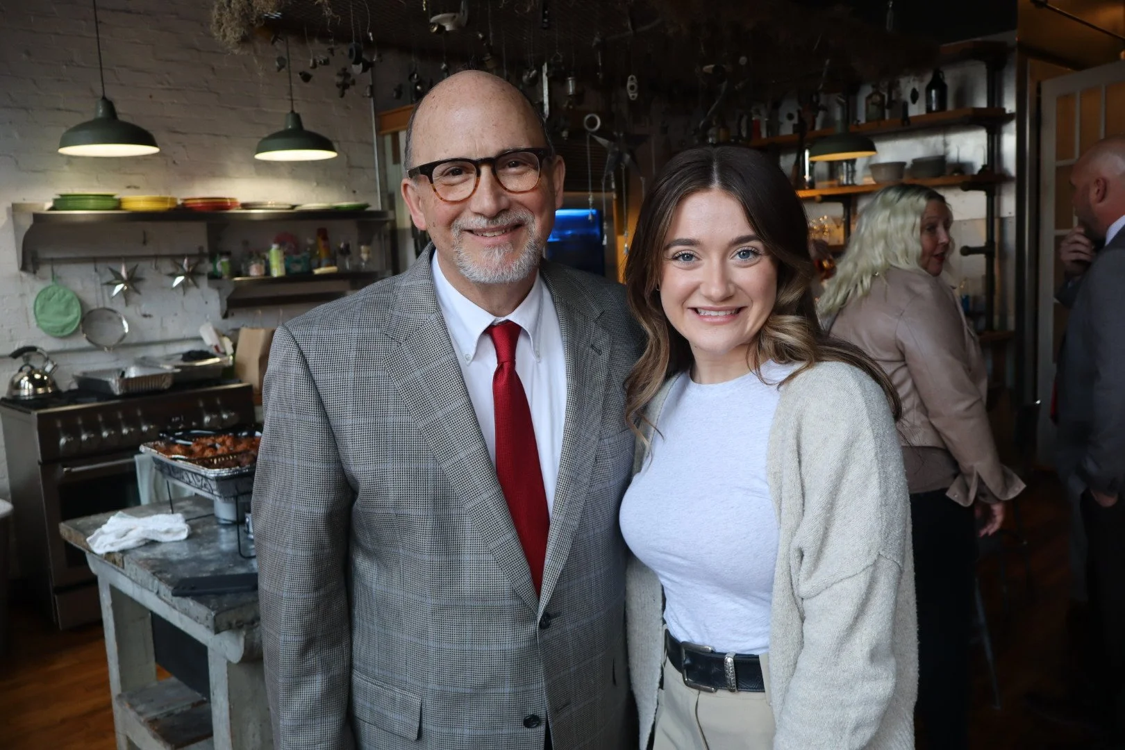 A smiling man in glasses and a woman with wavy brown hair pose together in a cozy, industrial-style kitchen with hanging lights, shelves, and a grill in the background.