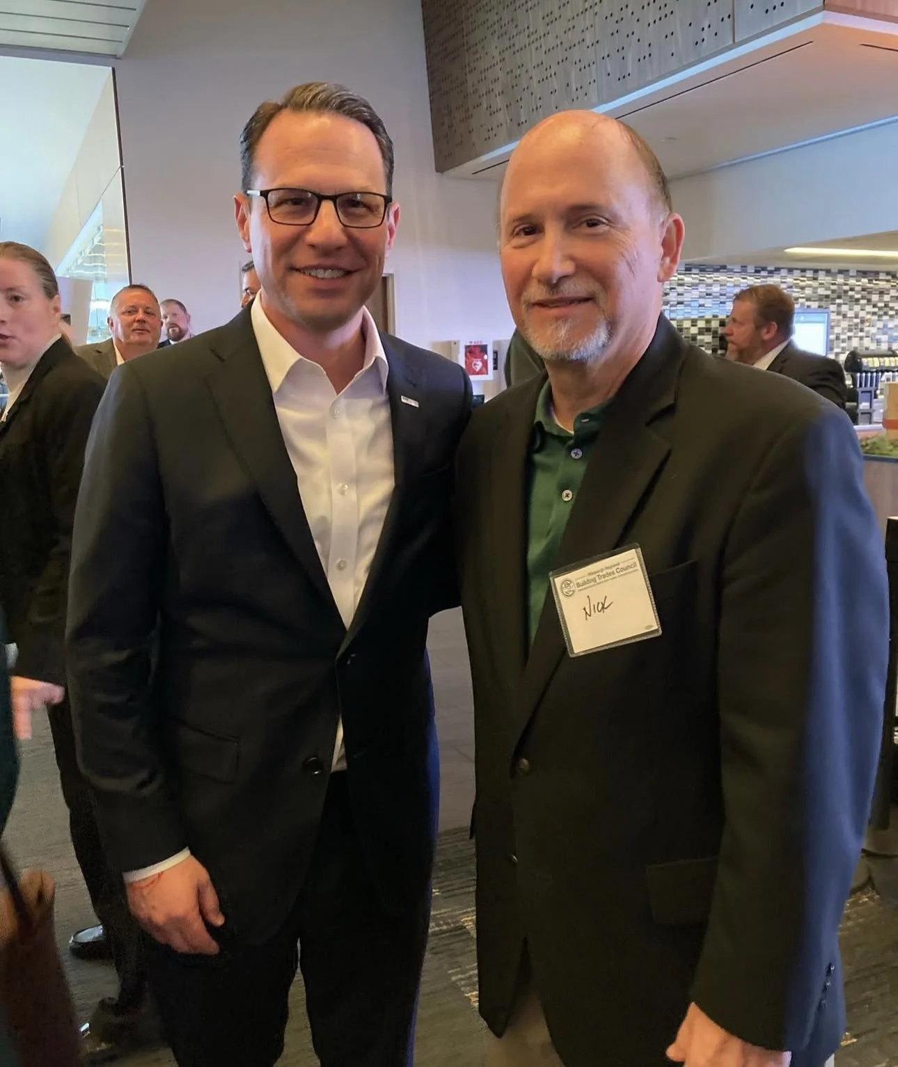 Two men in suits standing together at an indoor event, smiling at the camera. The man on the left has glasses and a white shirt, the man on the right has a beard, is wearing a green shirt, and has a name tag that says 'Nick'.