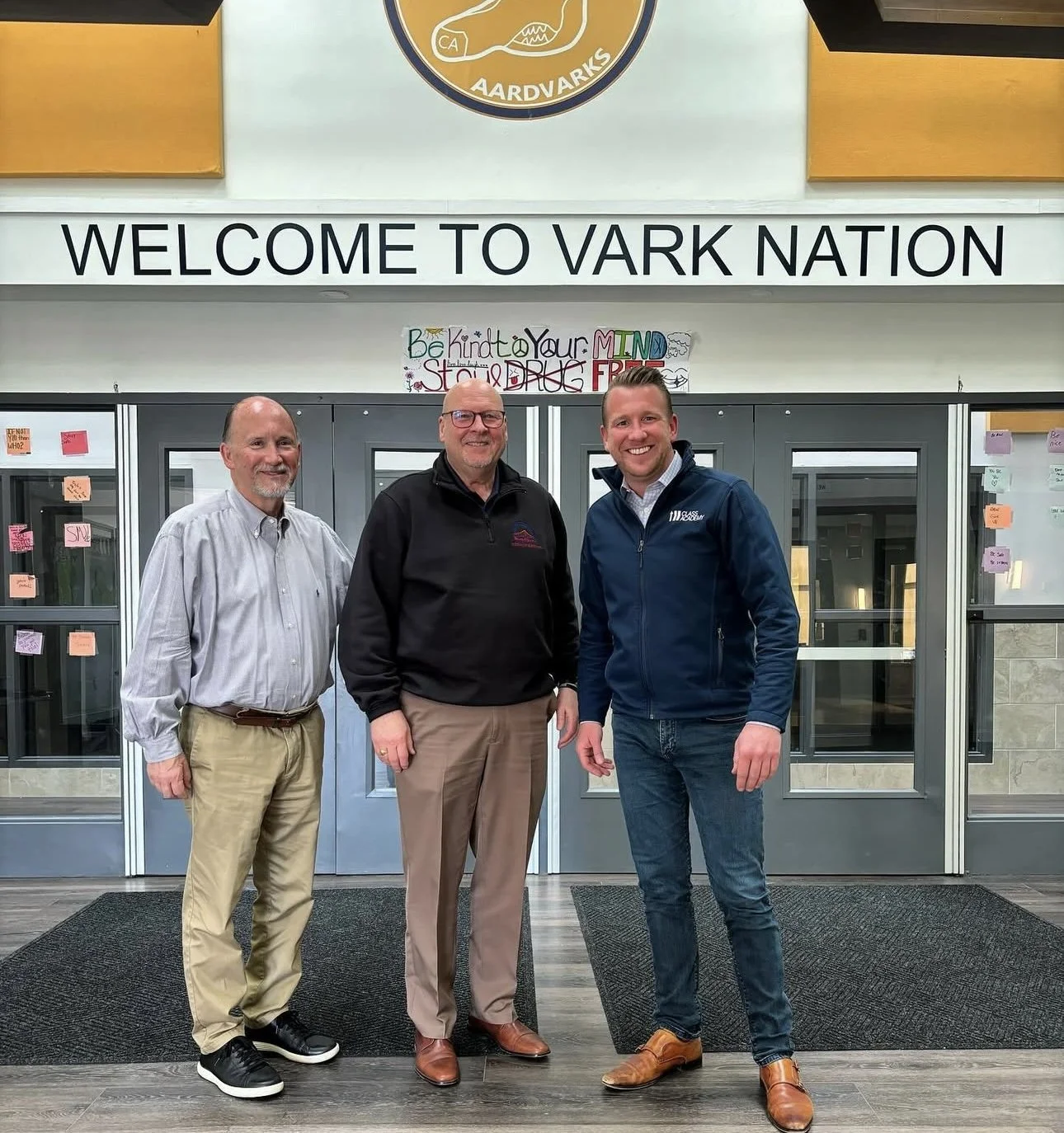 Three men standing inside a facility with signage that says "Welcome to Vark Nation" and a sign that reads "Be Kind to Your MTN D" in the background.