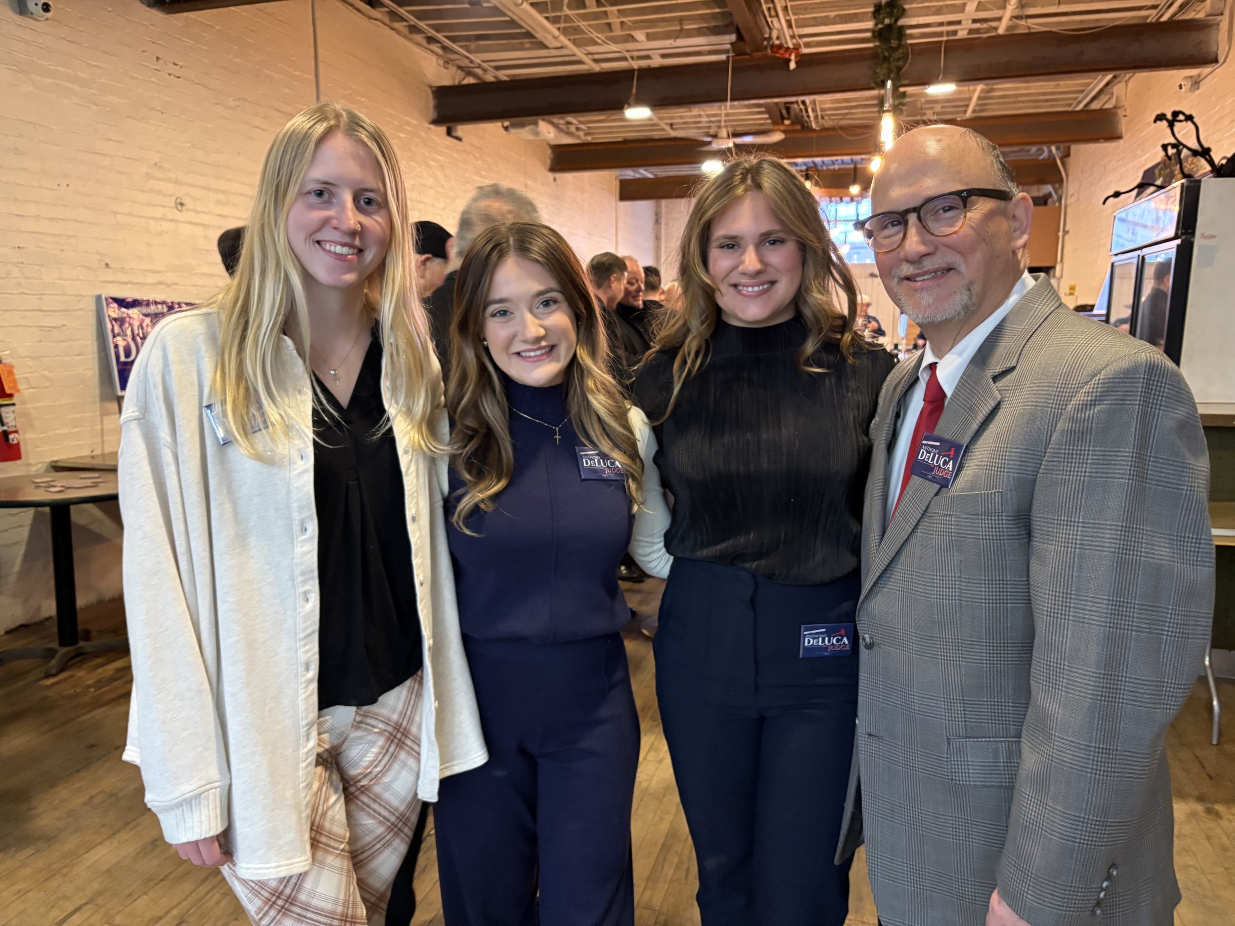 Four people standing together indoors at a social event, smiling for the camera, wearing name tags. The setting appears to be a casual or semi-formal gathering in a space with exposed brick walls and wooden beams.