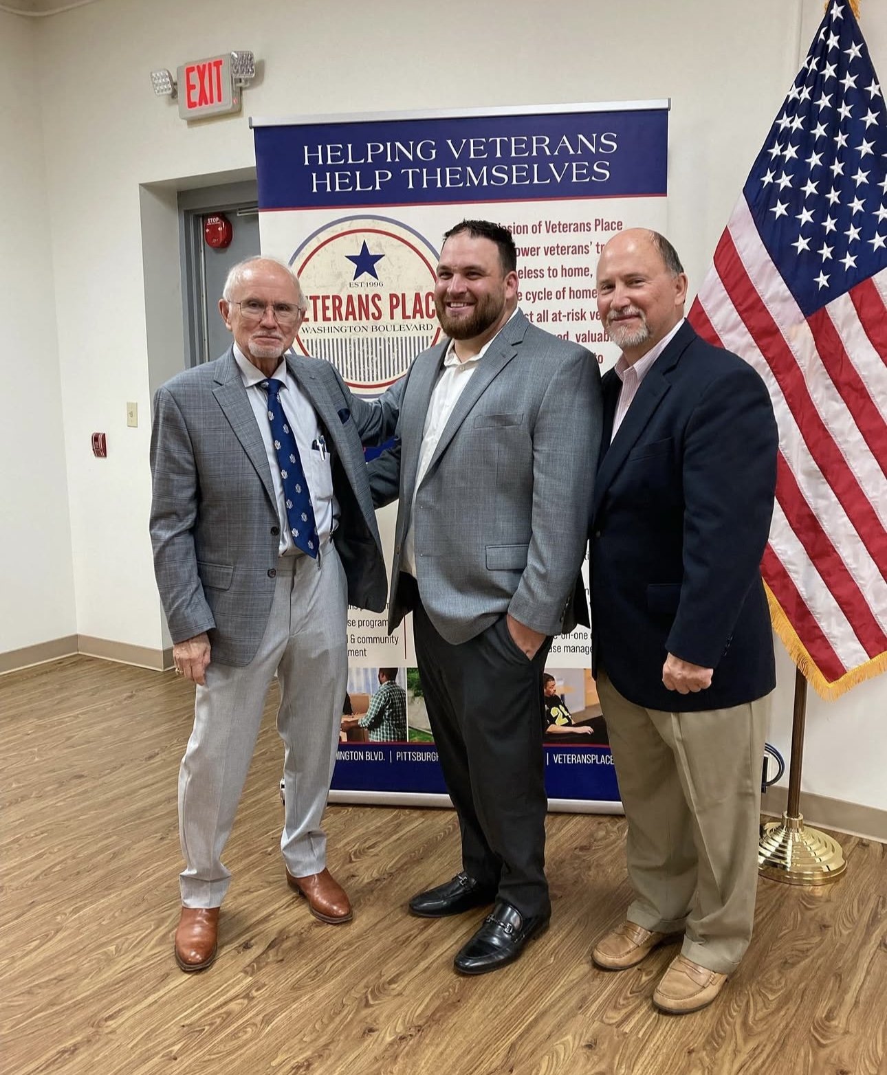 Three men in suits standing together indoors near a veterans' assistance sign, with an American flag on the right. The sign reads 'Helping Veterans Help Themselves' and includes the Veterans Place logo and information.