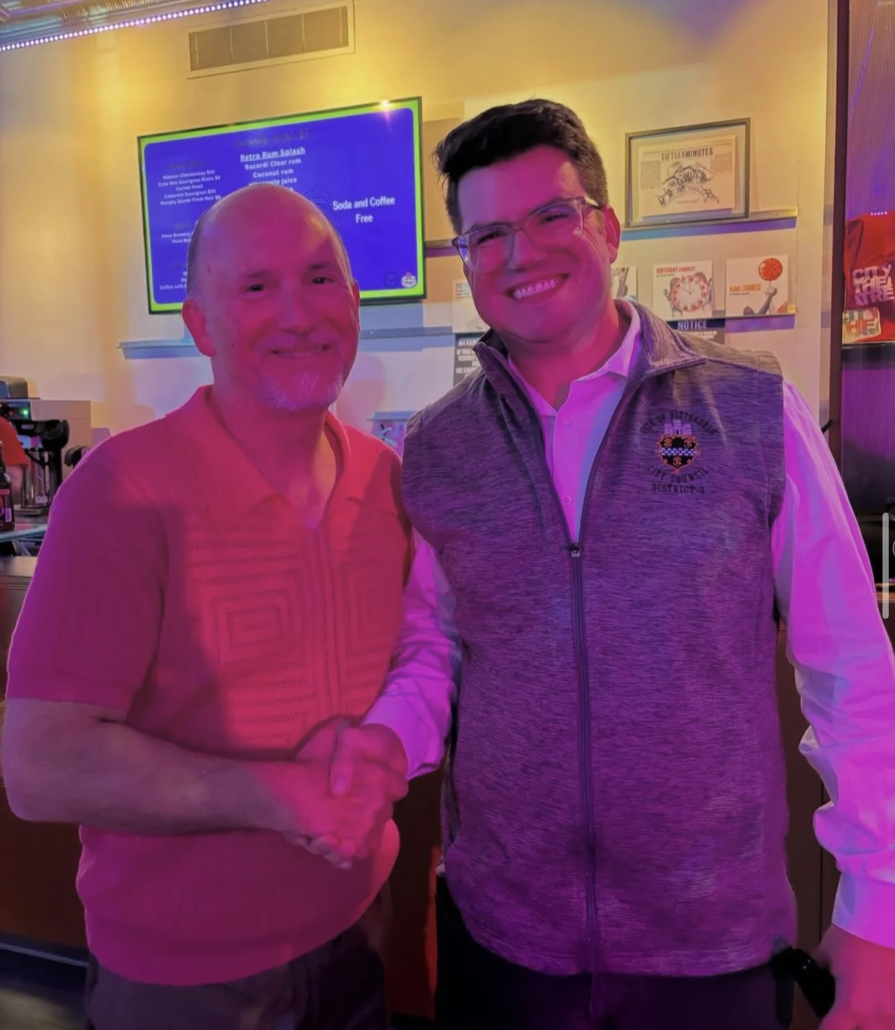 Two men shaking hands and smiling in a bar or restaurant with a colorful digital menu in the background.