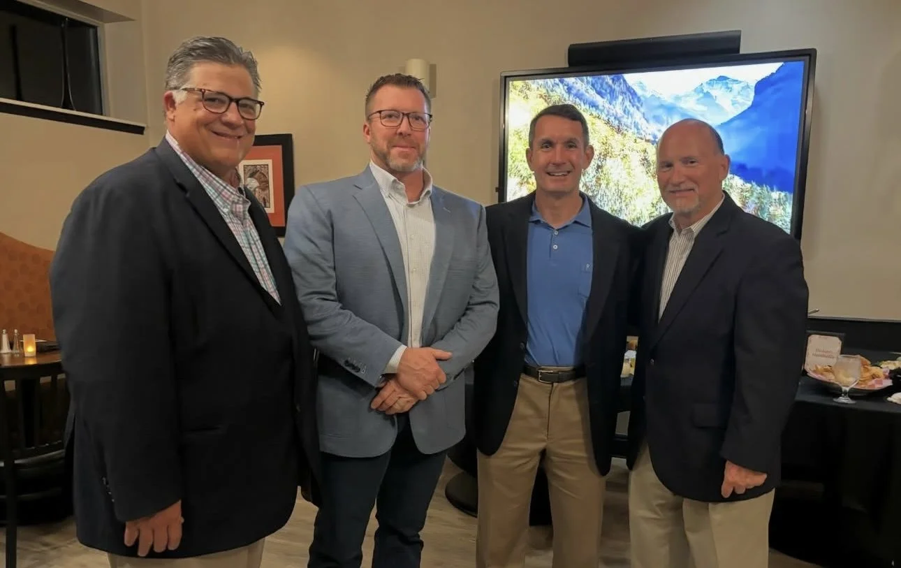 Four men dressed in business casual attire standing together in a room, with a large screen displaying a mountain landscape behind them.