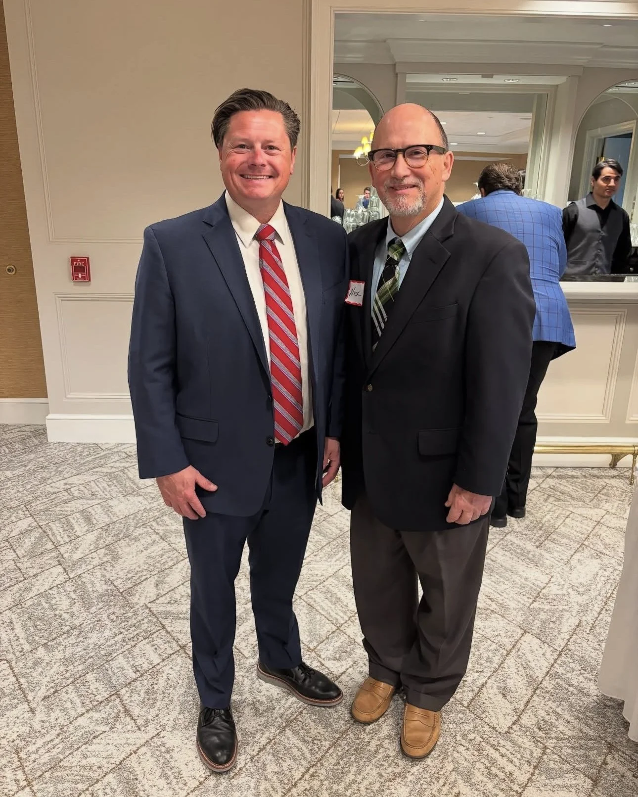 Two men dressed in suits smiling and posing for a photo in a formal setting, with a mirror showing other people in the background.