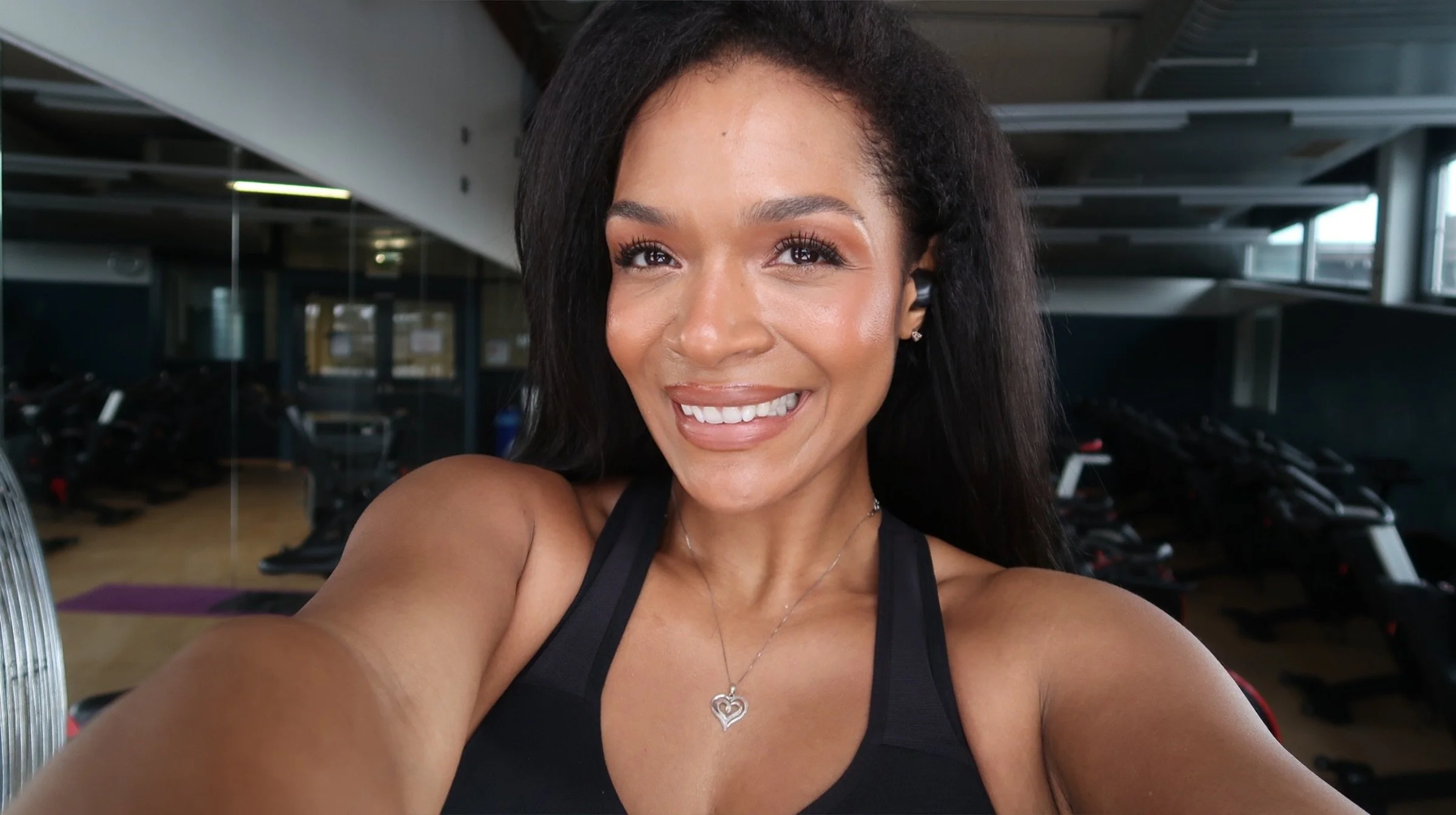 Eine strahlende junge Frau macht ein Selfie im Fitnessstudio, trägt ein schwarzes Sporttop und eine silberne Halskette, im Hintergrund sind Laufbänder und Fitnessgeräte sichtbar.