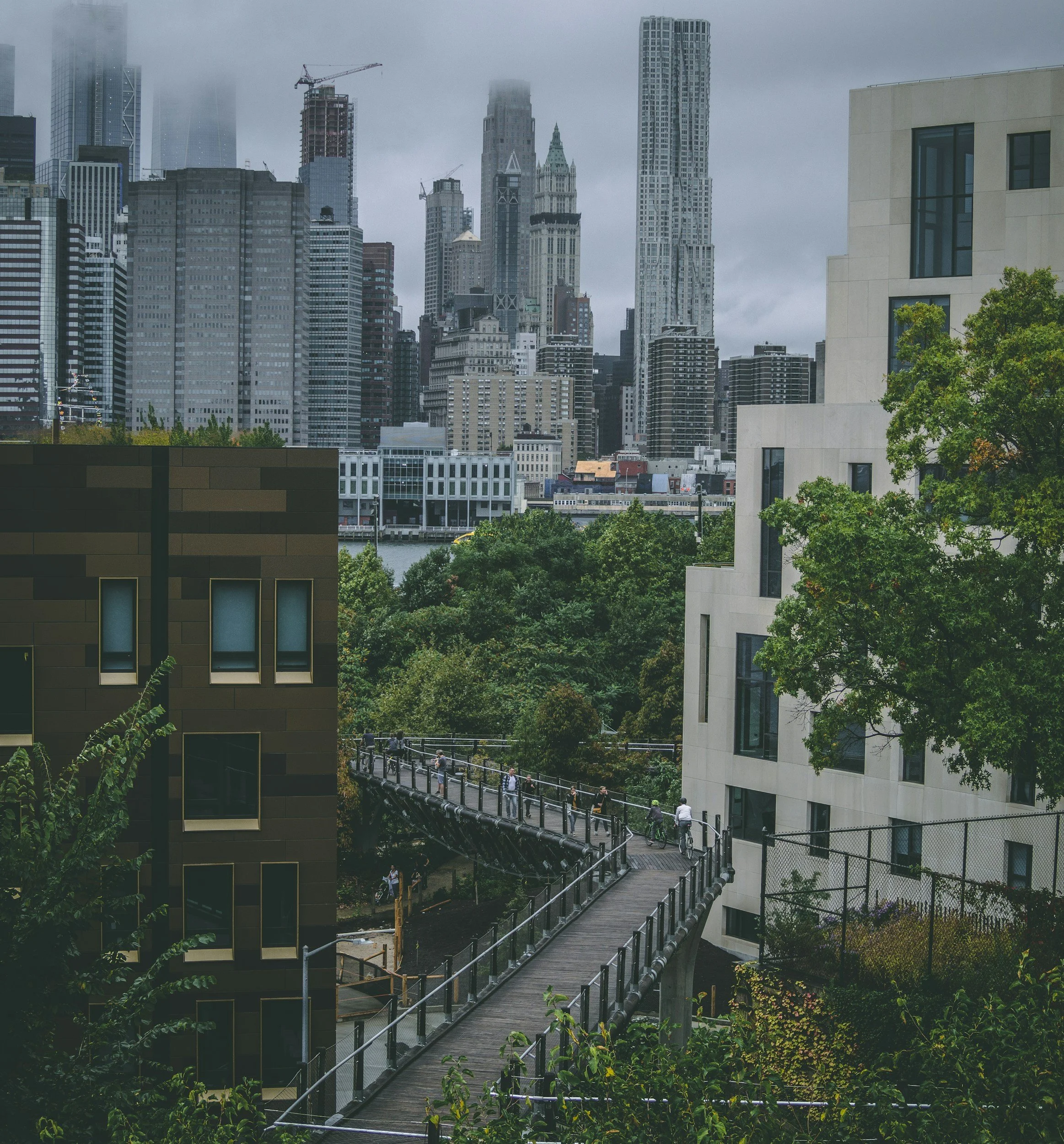Urban cityscape with skyscrapers in the background, a walking trail with people, trees, and modern buildings in the foreground, under cloudy skies.