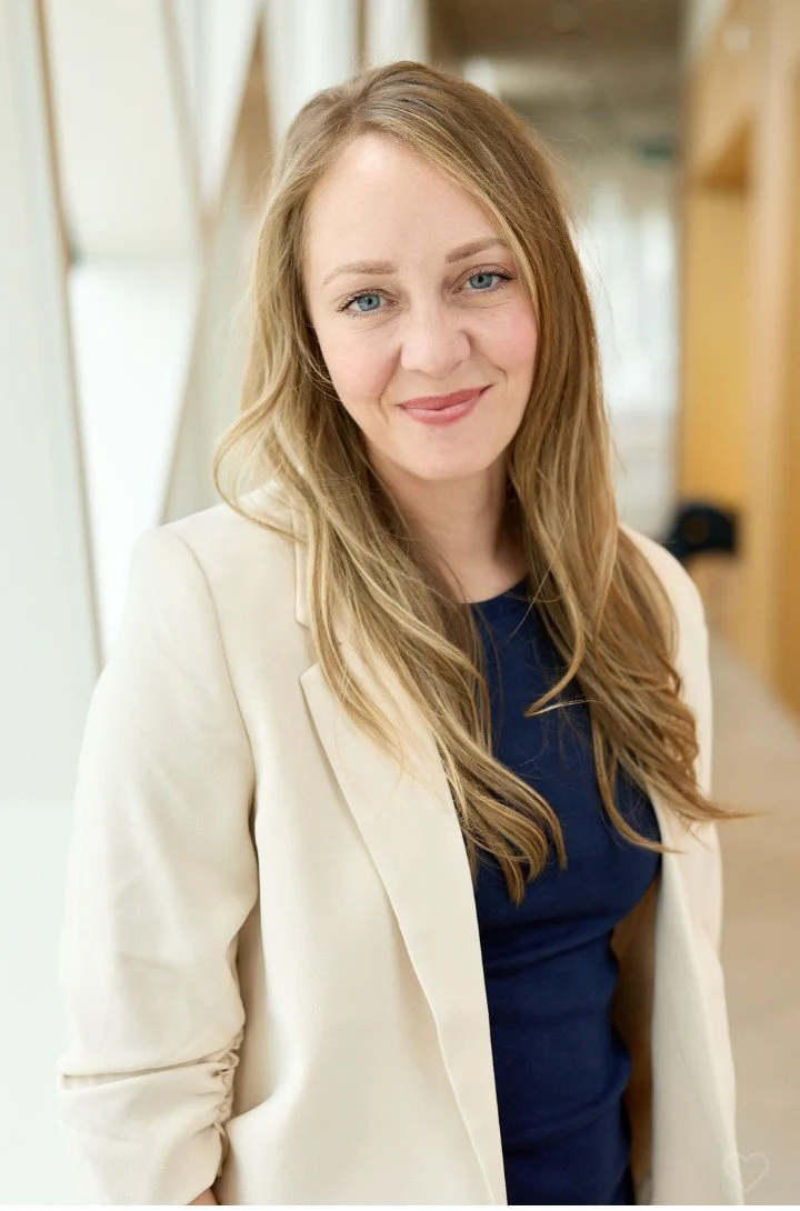 A woman with long, wavy blonde hair, light skin, and blue eyes smiling at the camera. She is wearing a cream-colored blazer over a navy blue top, standing in a well-lit indoor space.
