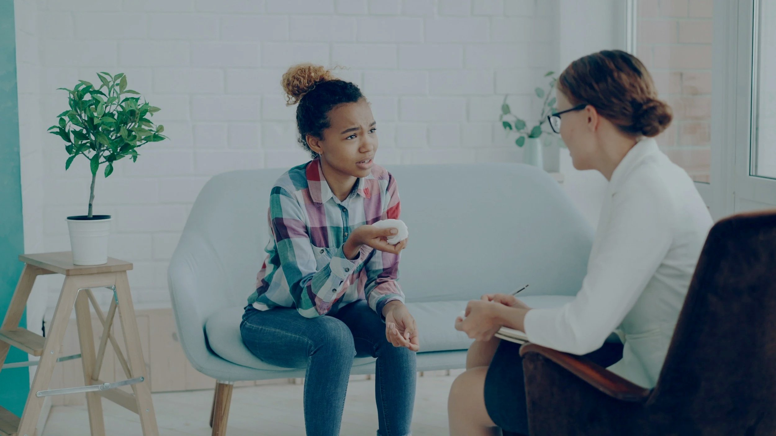 A young woman with curly hair and a plaid shirt sitting on a white couch holding a tissue, talking to a female healthcare professional with glasses and a white coat in a bright office with a potted plant on a wooden stand.