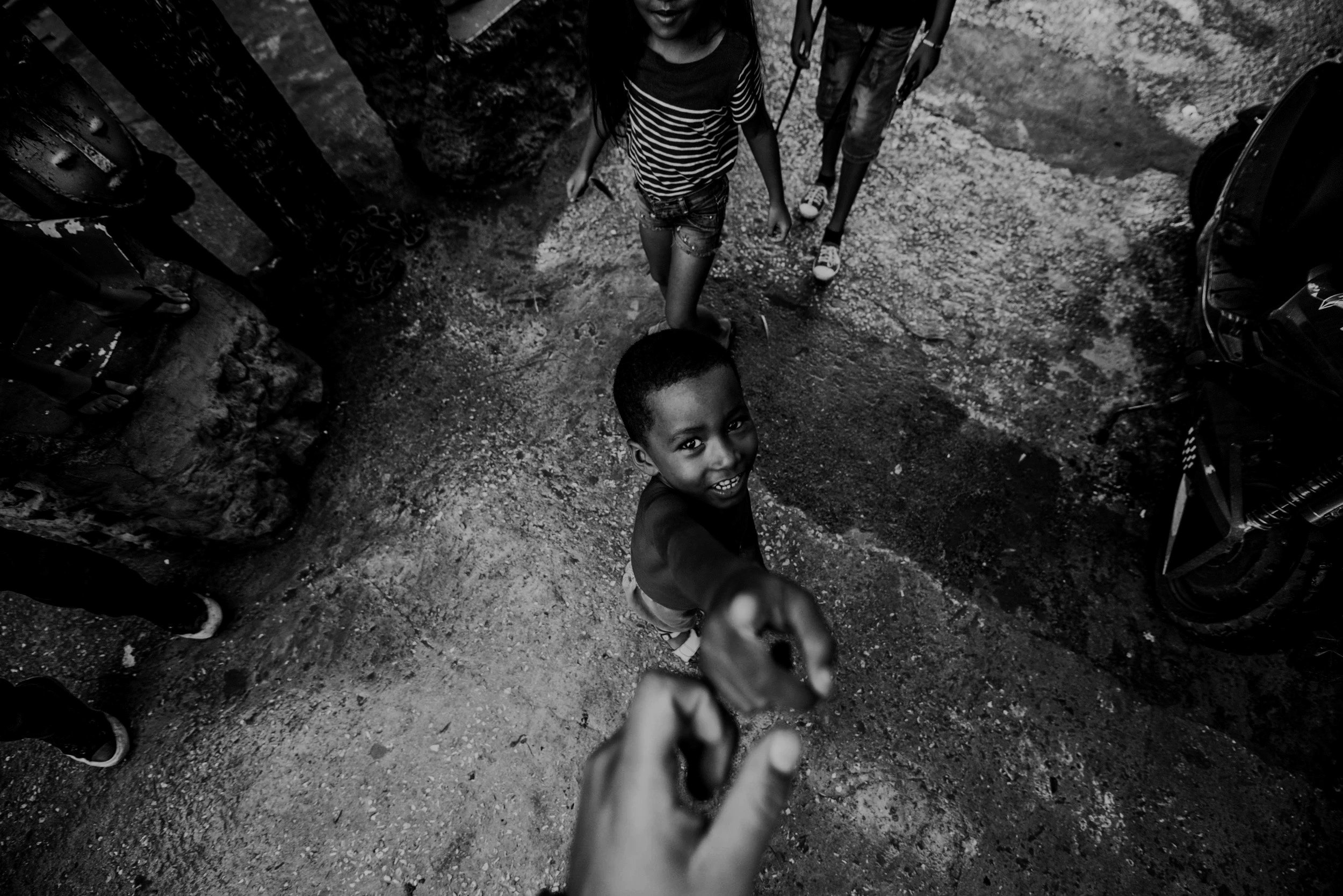 A black and white photograph of children walking on a dirt road, viewed from above. One child in the foreground is smiling at the camera, wearing a dark shirt. Other children are visible in the background, walking behind the smiling child.