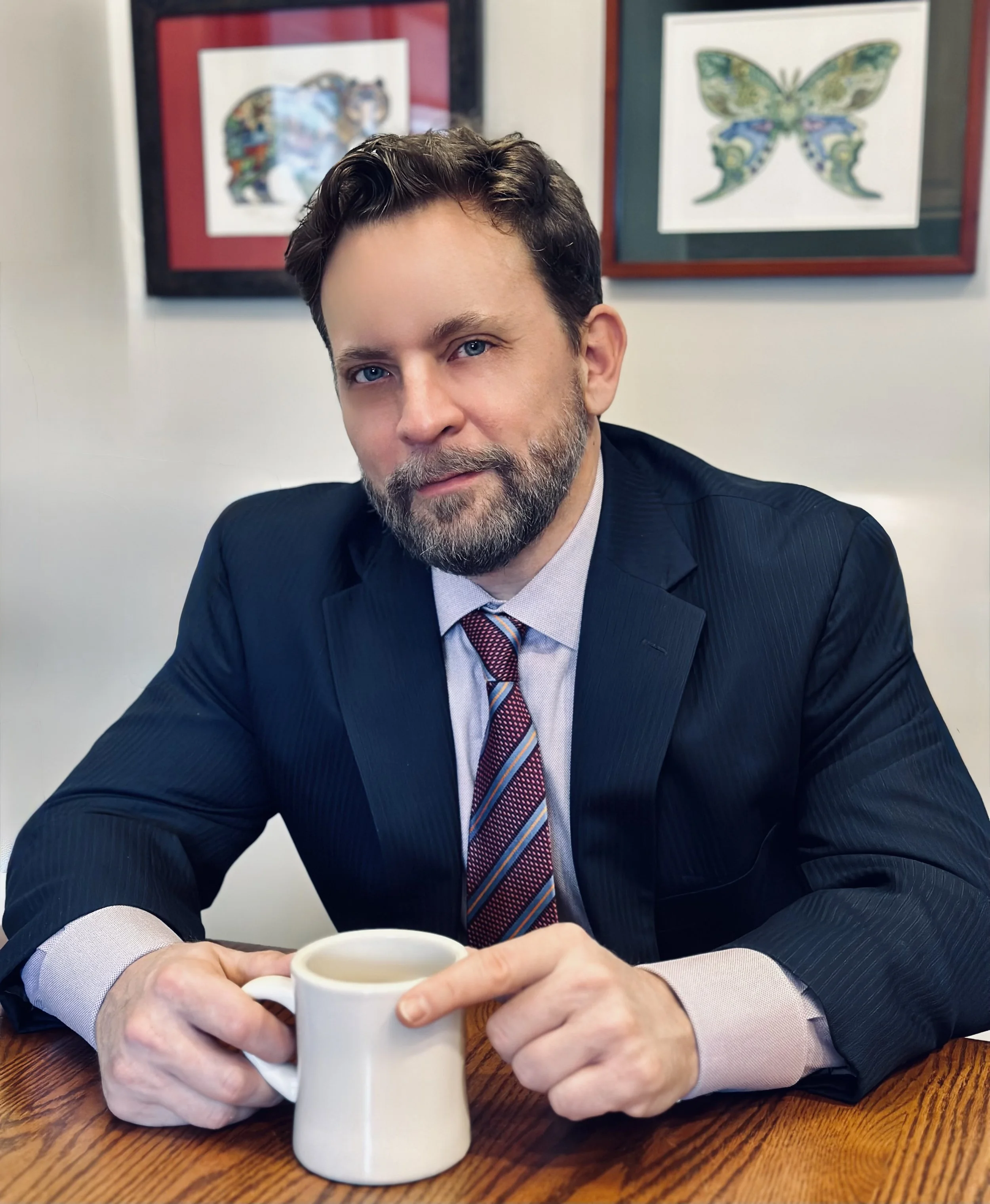 A man in a dark suit, light shirt, and colorful striped tie sitting at a wooden table, holding a white coffee mug. Behind him are framed butterfly and animal artwork on the wall.