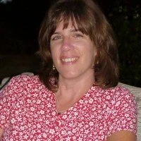 Woman with shoulder-length brown hair wearing a pink and white floral blouse, smiling outdoors at night.