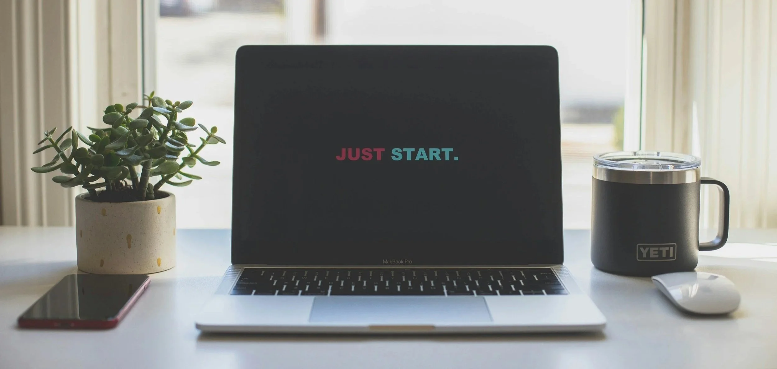 A laptop on a desk with the words "JUST START." on its screen, a potted succulent plant on the left, a smartphone on the far left, a Yeti mug with a clear lid and a mouse on the right, and a window with curtains in the background.