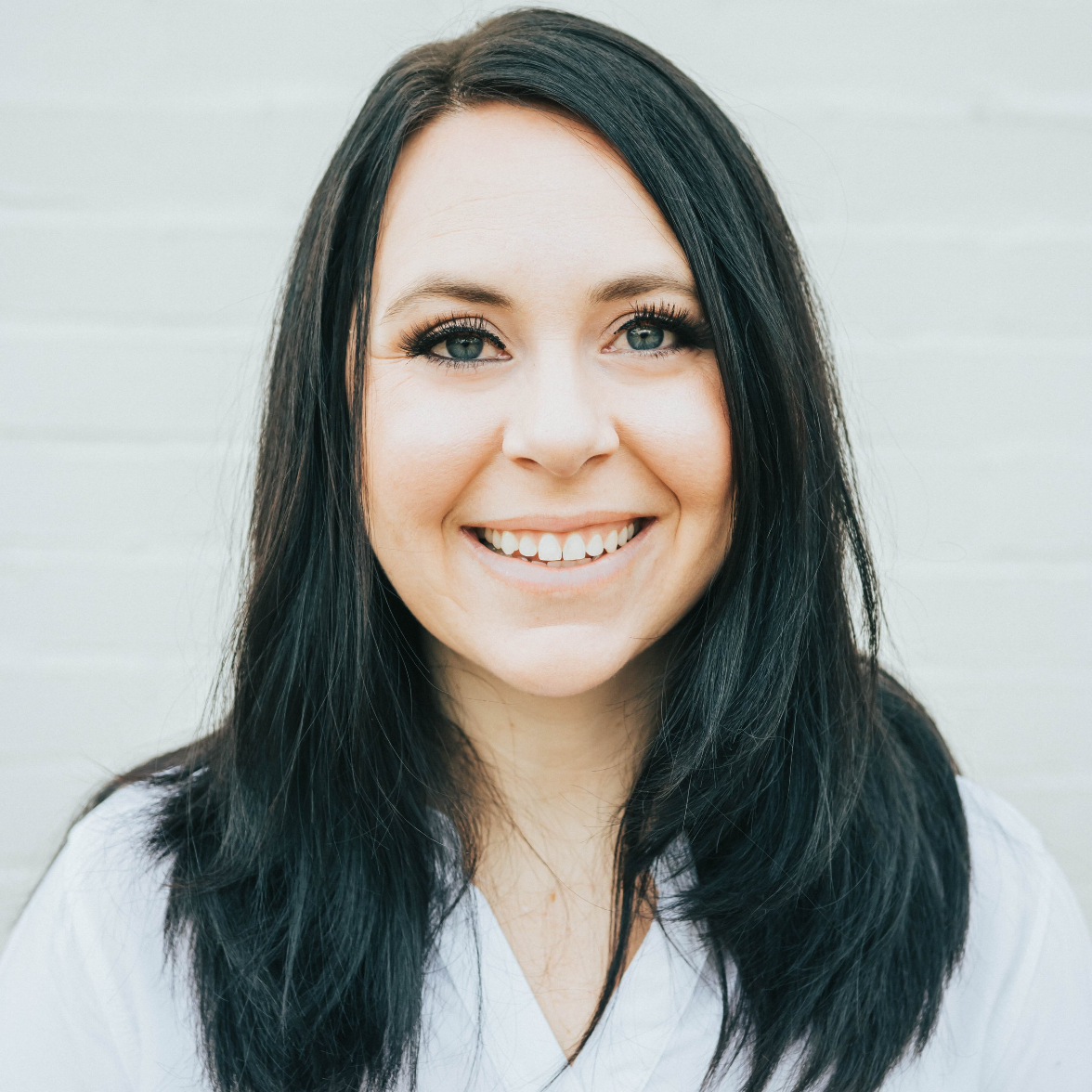 Close-up of a woman with long black hair, blue eyes, and a bright smile, wearing a white shirt, against a light background.