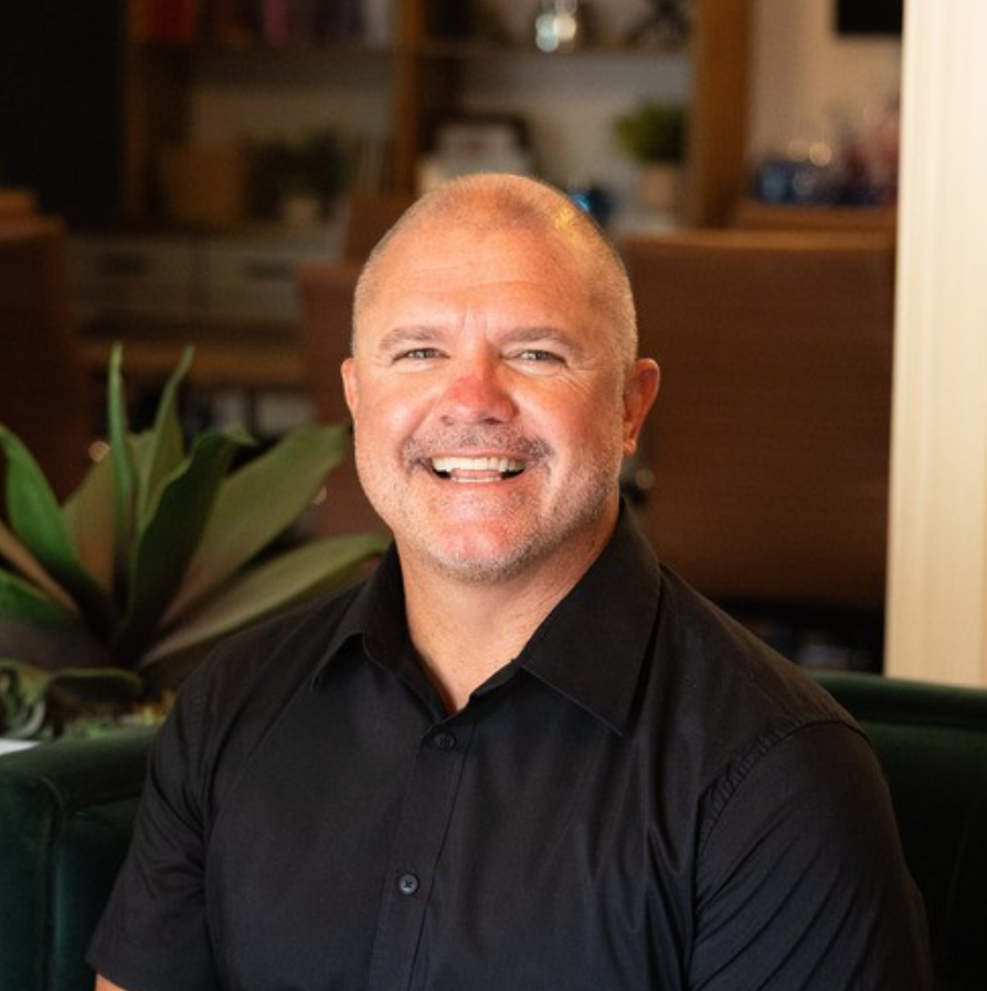 A smiling man with a bald head and facial hair, wearing a black shirt, sitting in a cozy, well-lit indoor setting with plants and furniture in the background.