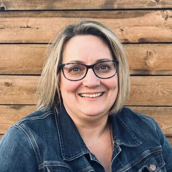 A woman with blonde hair and glasses smiling, wearing a denim jacket, in front of a wooden wall.
