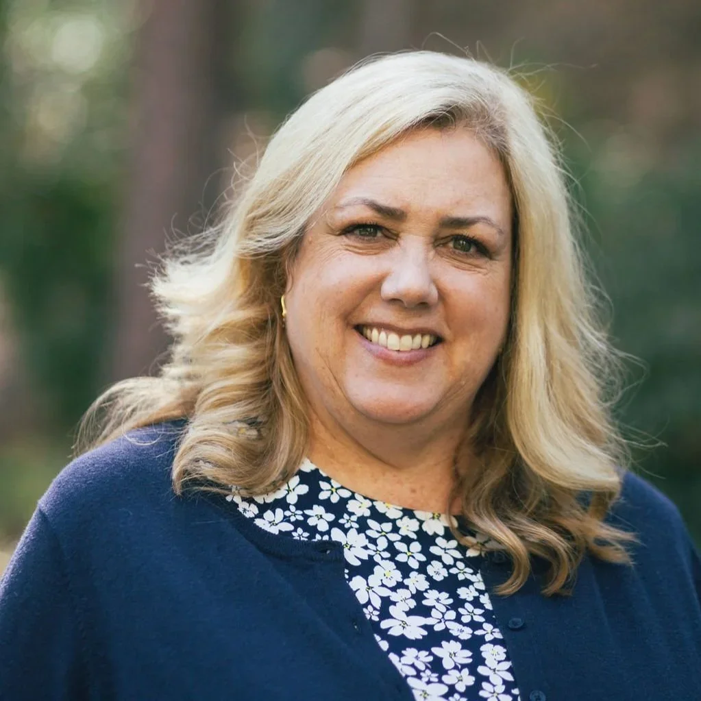 Portrait of a woman with blonde hair smiling outdoors, wearing a navy blue top with a floral pattern and a dark blazer.