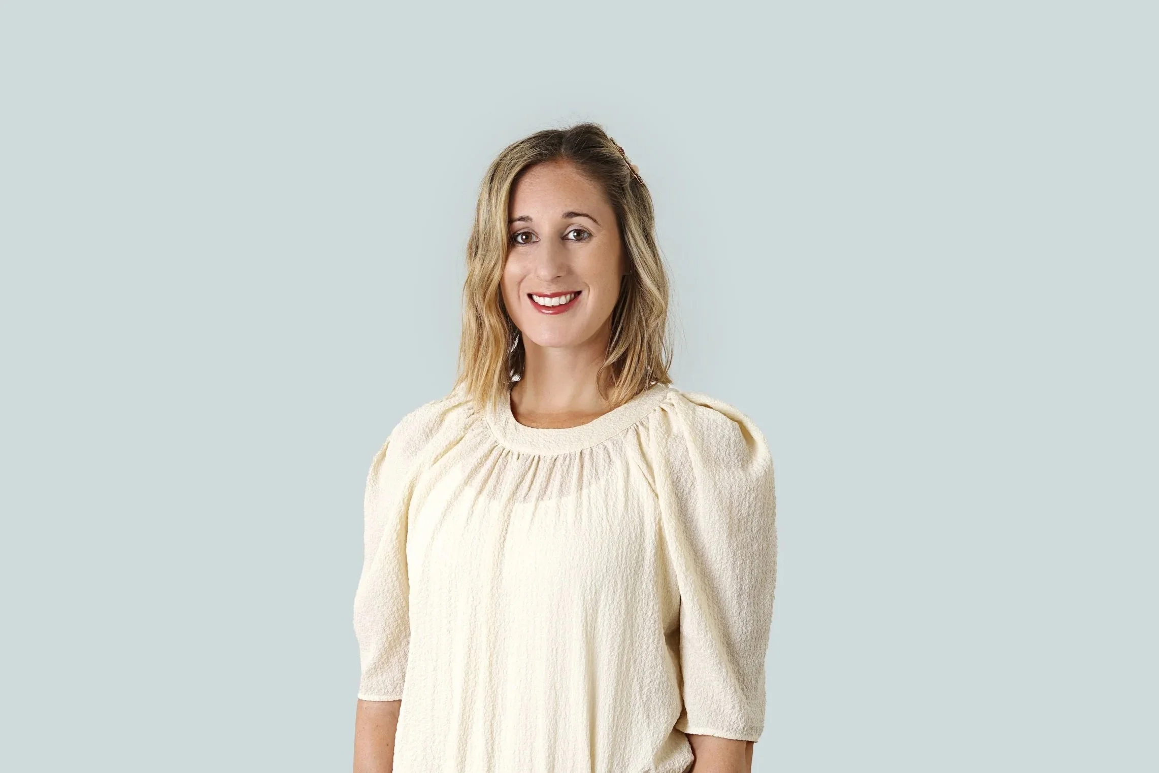 A woman with shoulder-length blonde hair wearing a cream-colored blouse smiling at the camera against a plain light background.