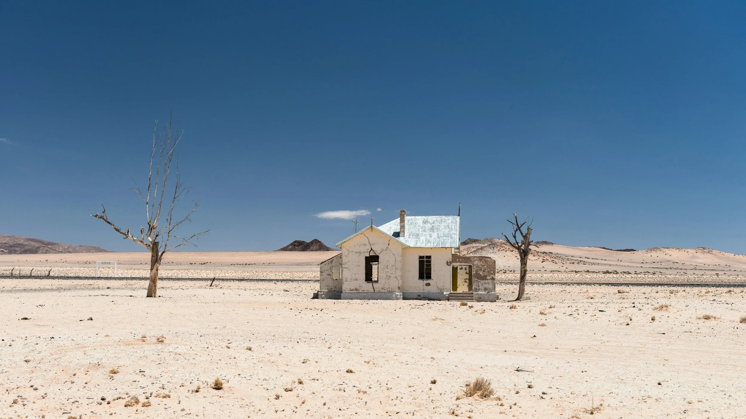 Abandoned house with white walls and a metal roof in a barren desert landscape, flanked by two leafless trees under a clear blue sky.