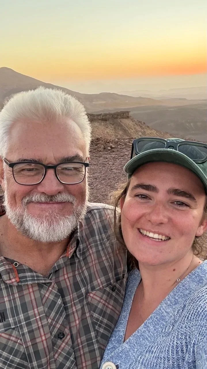 A smiling older man with white hair, glasses, and a beard taking a selfie with a woman with long hair wearing a cap, against a desert landscape at sunset.