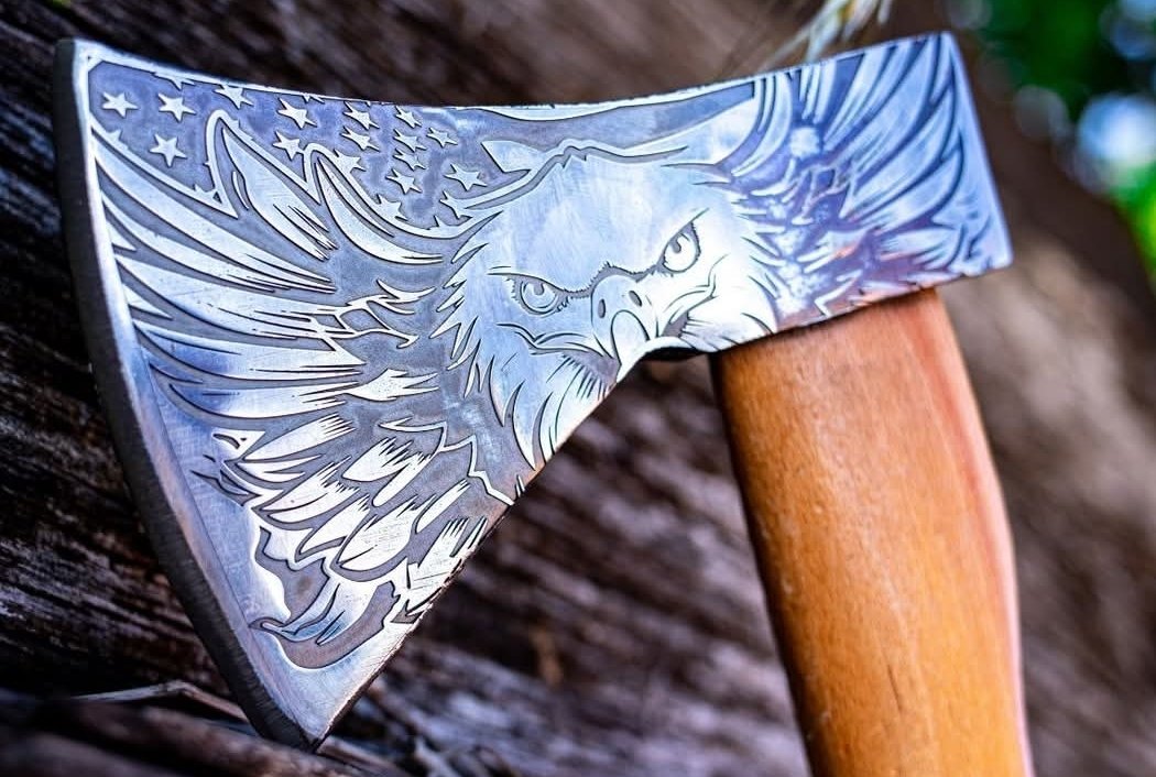 Close-up of a hatchet with an engraved eagle design on the metal blade and a wooden handle, leaning against a rough wooden surface.