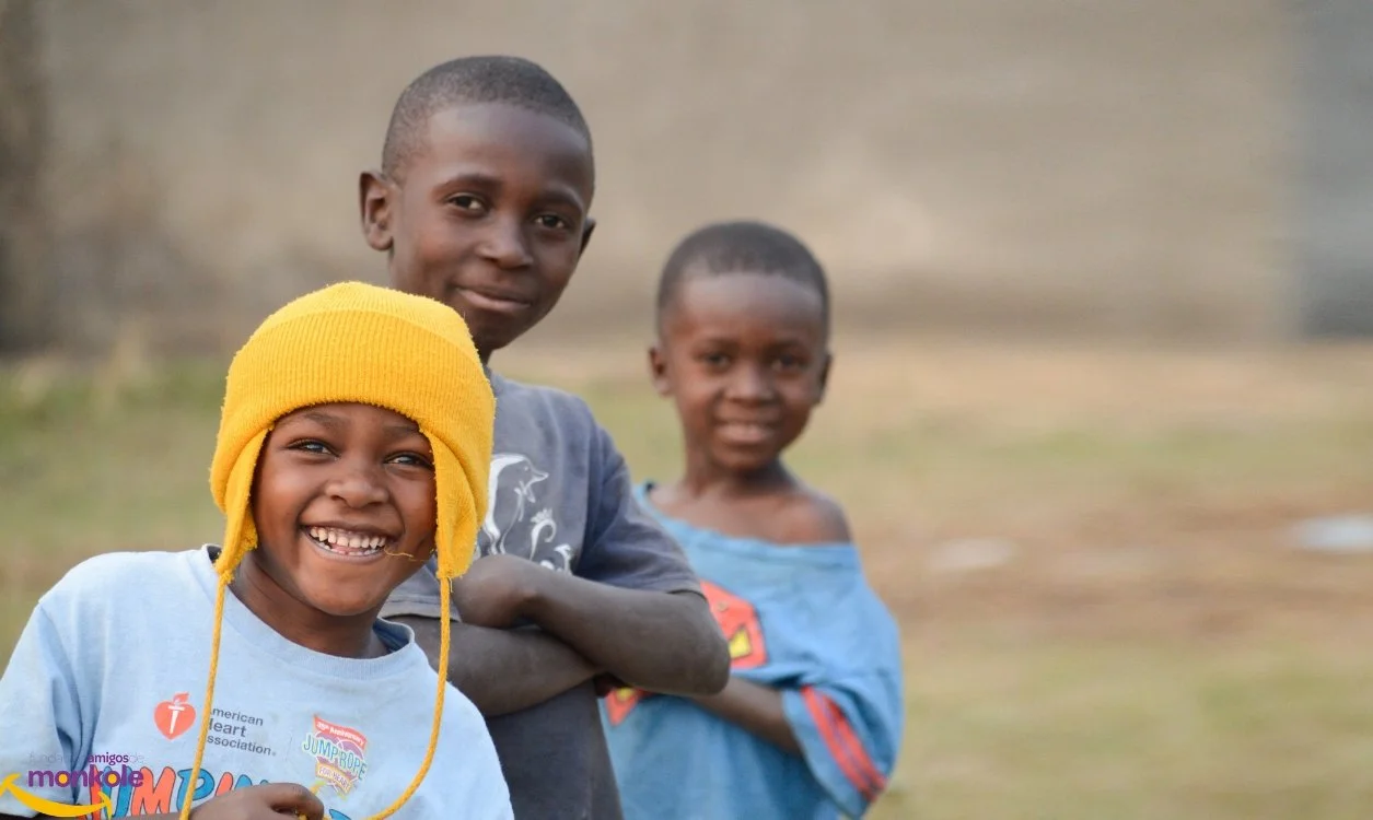 Three smiling children outdoors, one wearing a yellow hat, the other two with short hair, standing on a grassy area.