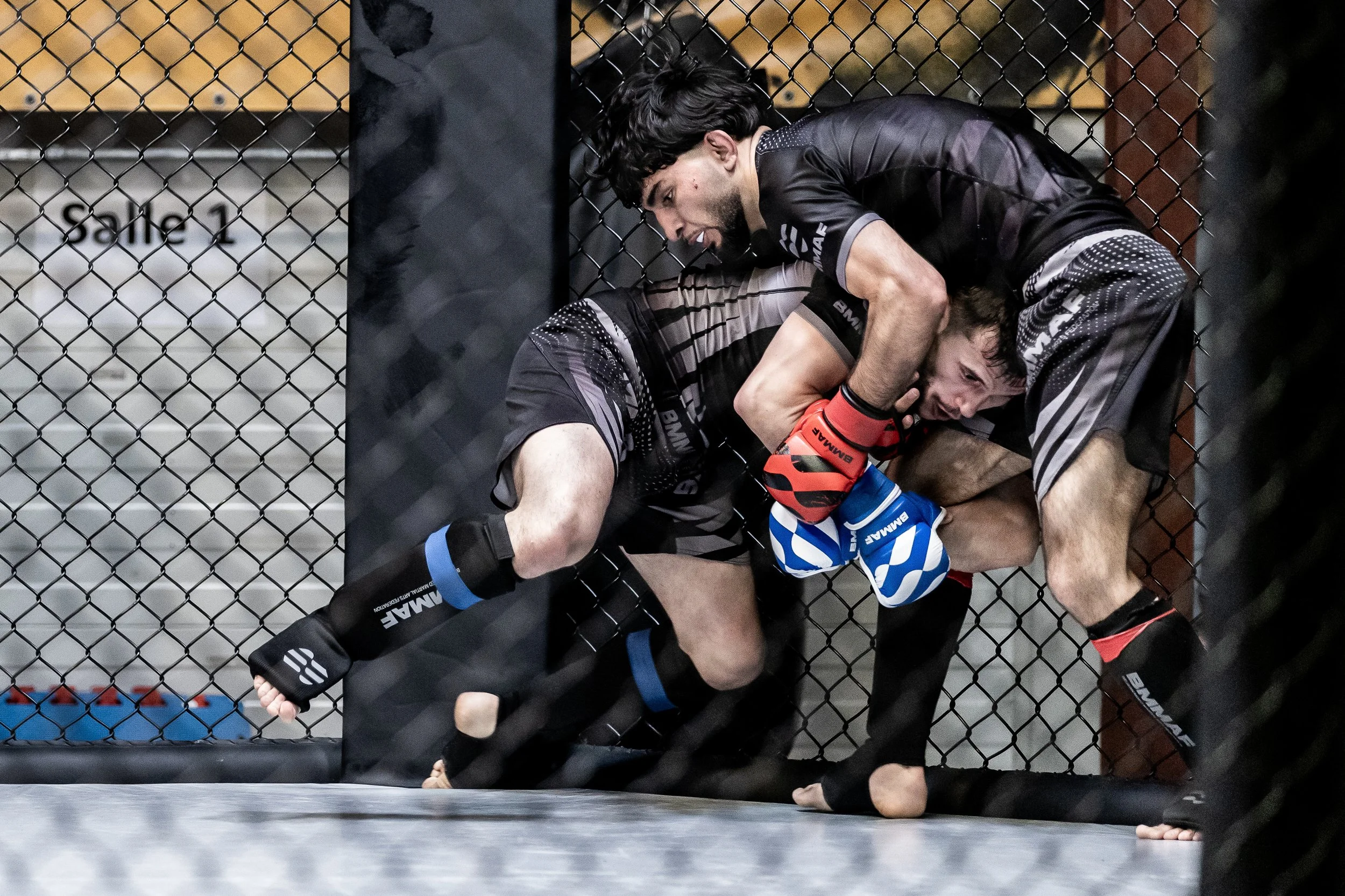 Twee mannen vechten binnen en om een metalen kooi, een op de grond, de ander erboven, beide gekleed in gevechtsshirts en handschoenen in een MMA-gevecht.