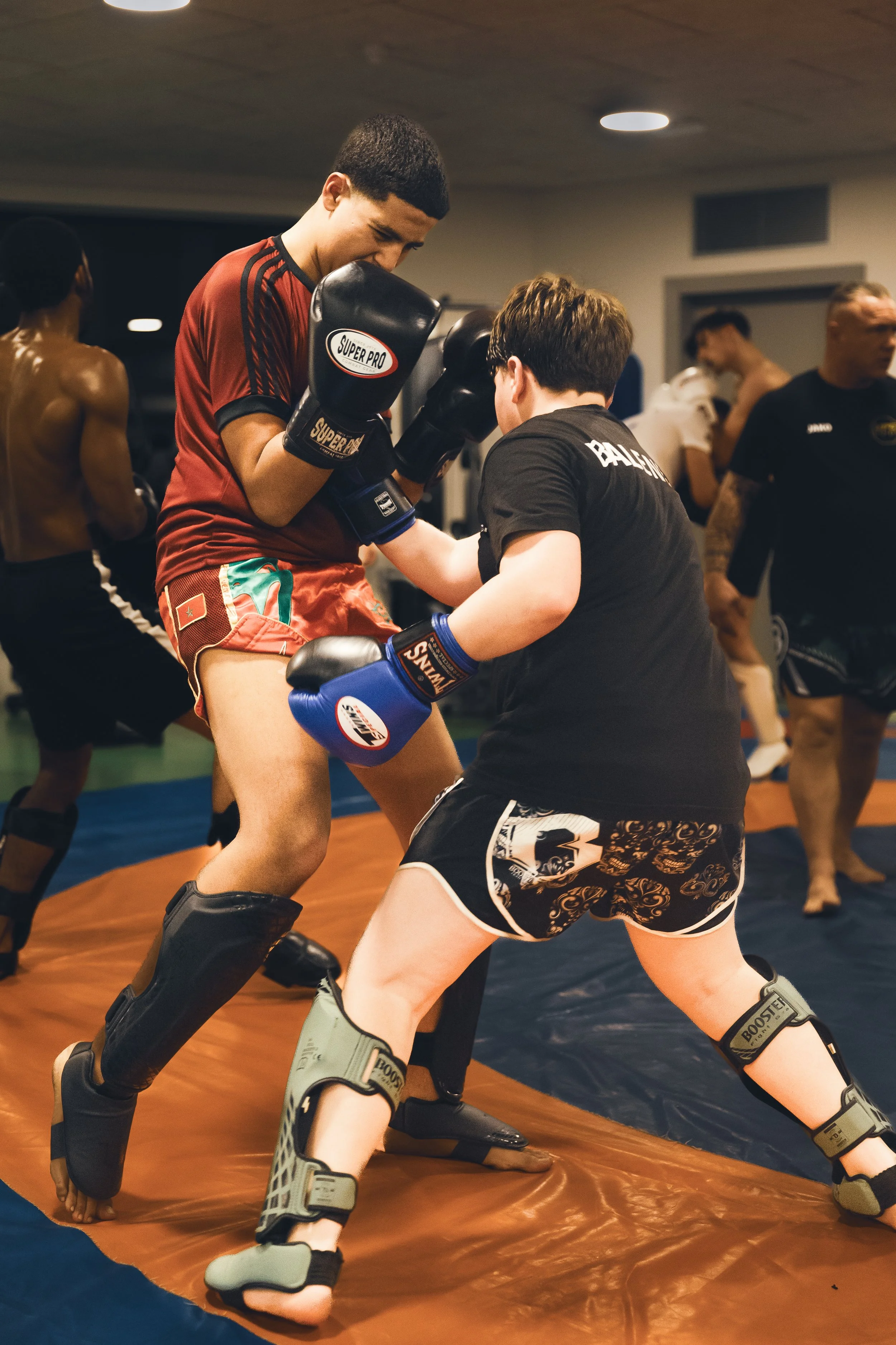 Twee mannen trainen in een Muay Thai gym, met een groep mensen op de achtergrond, de man aan de linkerkant heeft blauwe kickbokshandschoenen en zit in een vechthouding, de andere man ondersteunt hem met een stoot, beiden dragen sportkleding.