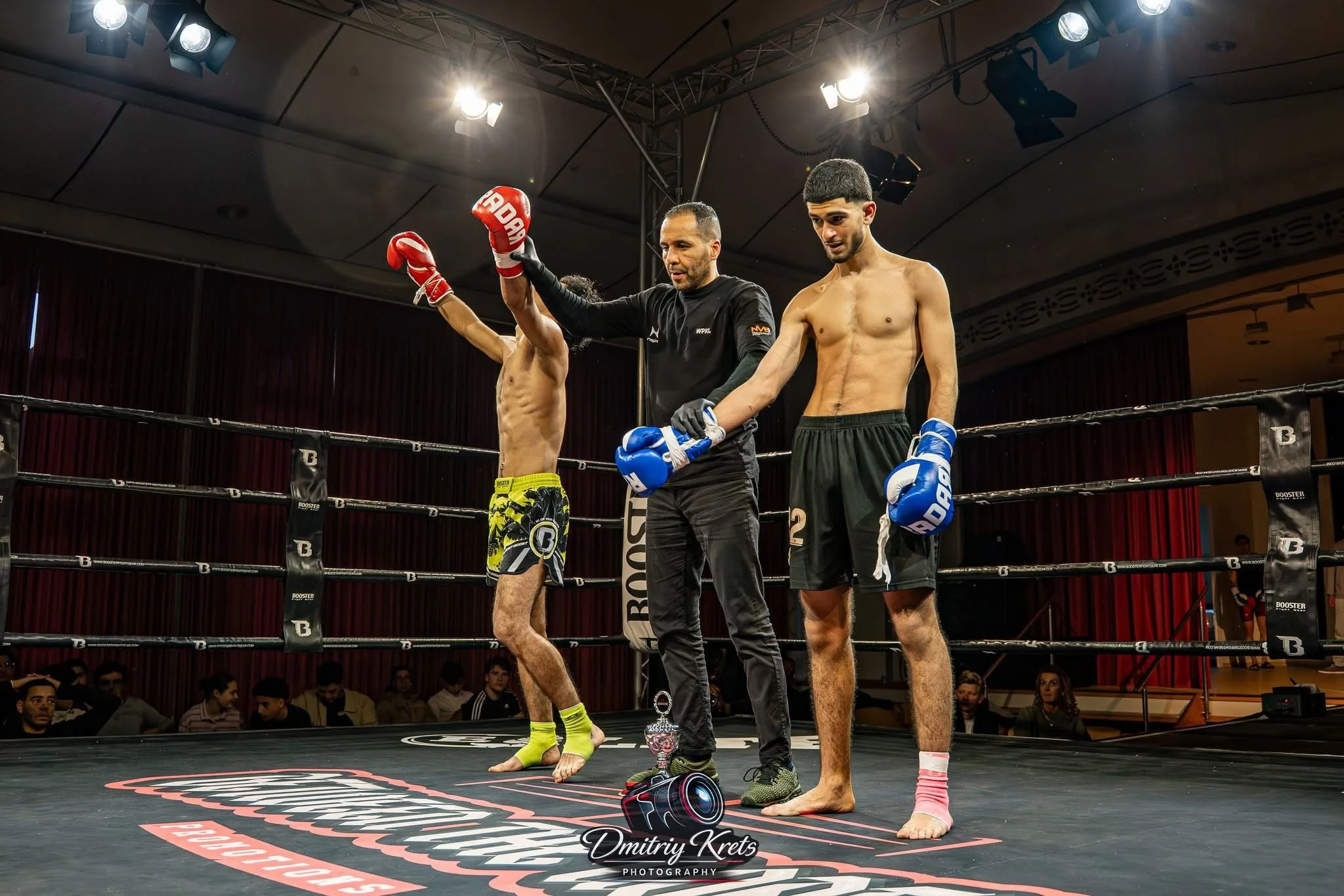 Twee vechters in een ring na een wedstrijd, een winnaar en een verliezer, met de winnaars hand omhoog gehouden. Mensen kijken toe en applaudisseren op de achtergrond.
