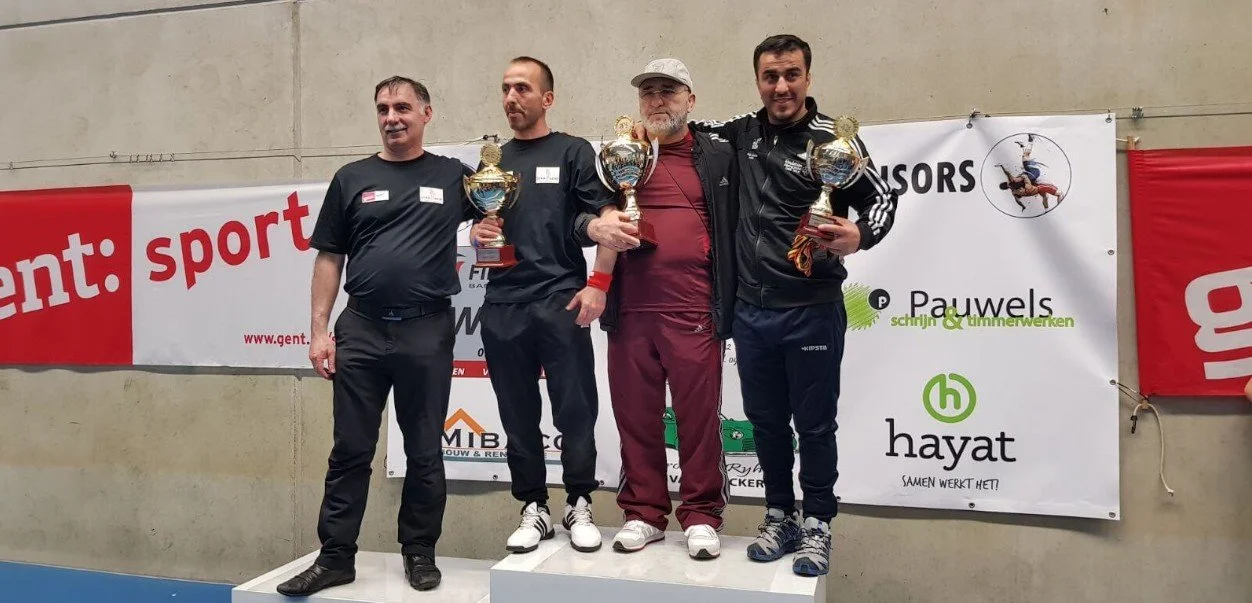 Vier mannen op een prijsuitslagpodium, drie van hen houden trofeeën en vieren hun overwinning. Ze zijn in een sporthal met banners op de achtergrond.