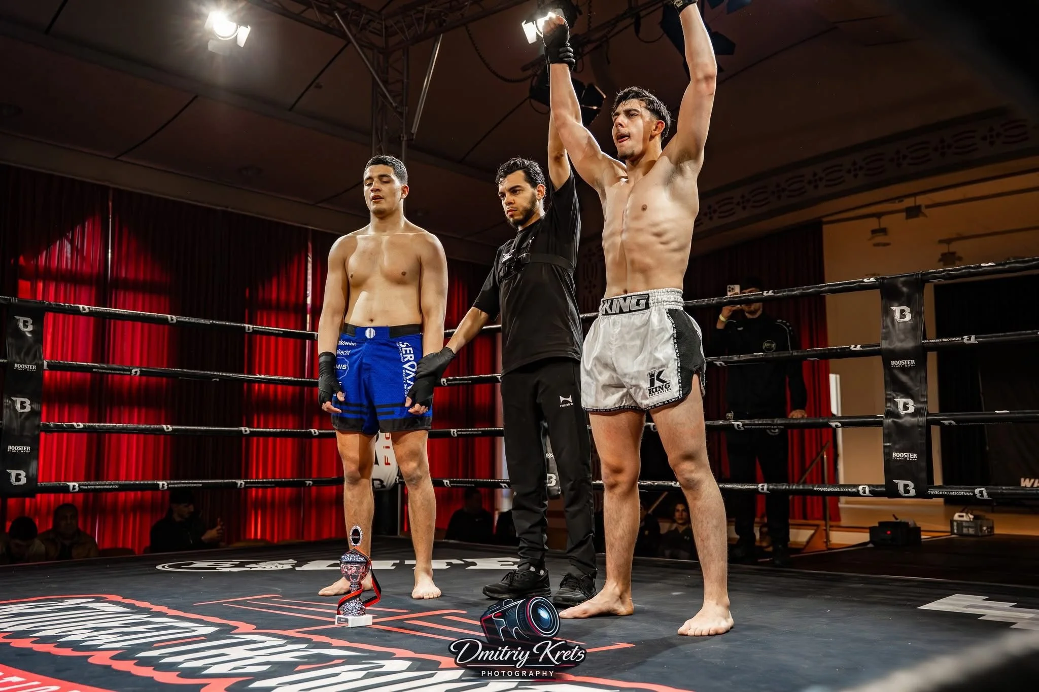 Twee kickboksers staan in de ring met een scheidsrechter die hun overwinning bevestigt, een van hen met omhoog geslagen armen. Een trofee ligt voor hen op de mat.
