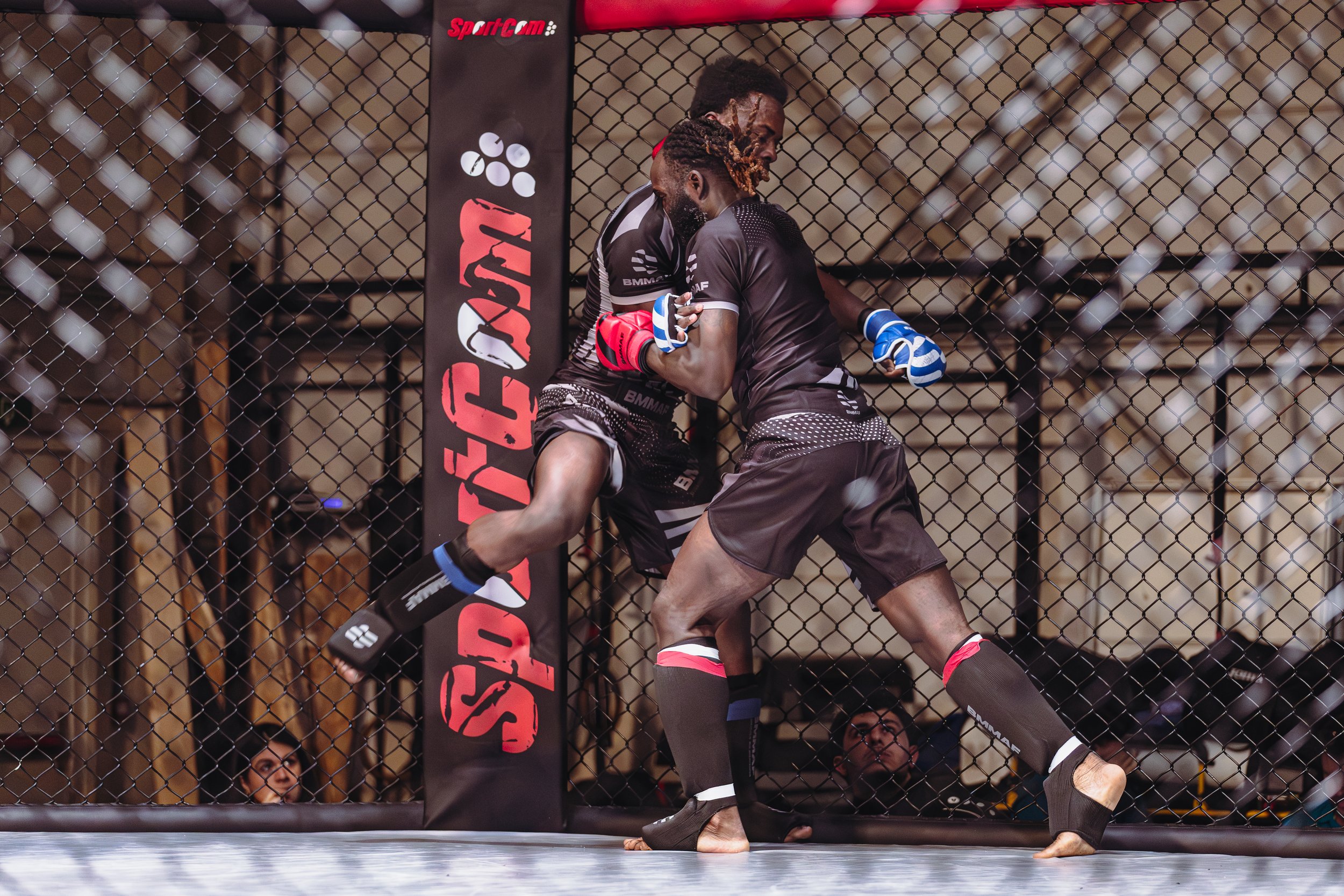 Twee jonge mannen in vechtsportoutfits die in een mixed martial arts cage vechten, terwijl een vrouw toekijkt en de scene door het hek wordt gefilmd.