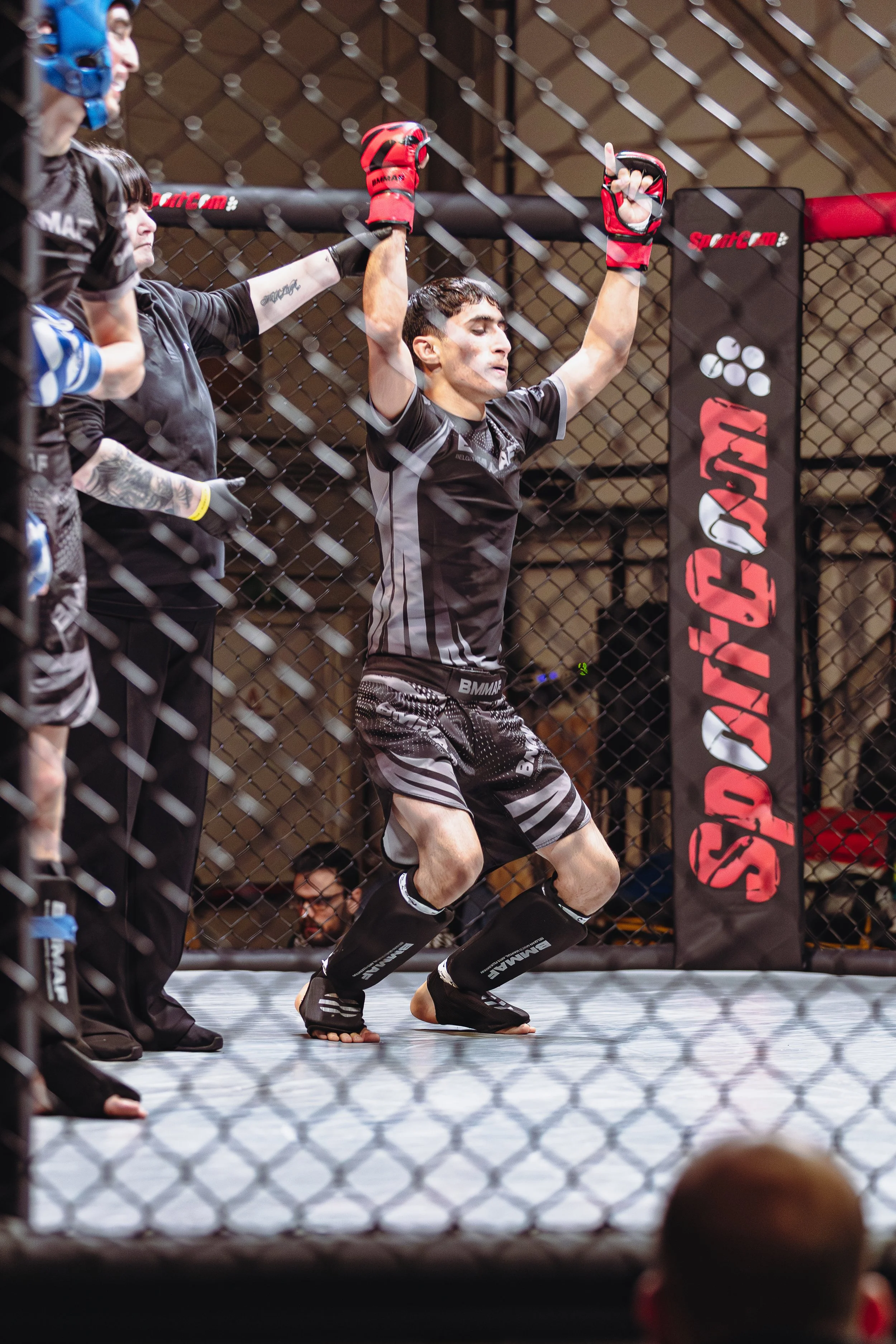 Een man in een MMA-ring, met een gevechtshandschoen in de hand en gekleed in zwart en grijs, lijkt klaar te zijn om te vechten. Hij staat tegen de muur van het rooster, terwijl officials en andere deelnemers aan de zijkant van de ring staan.