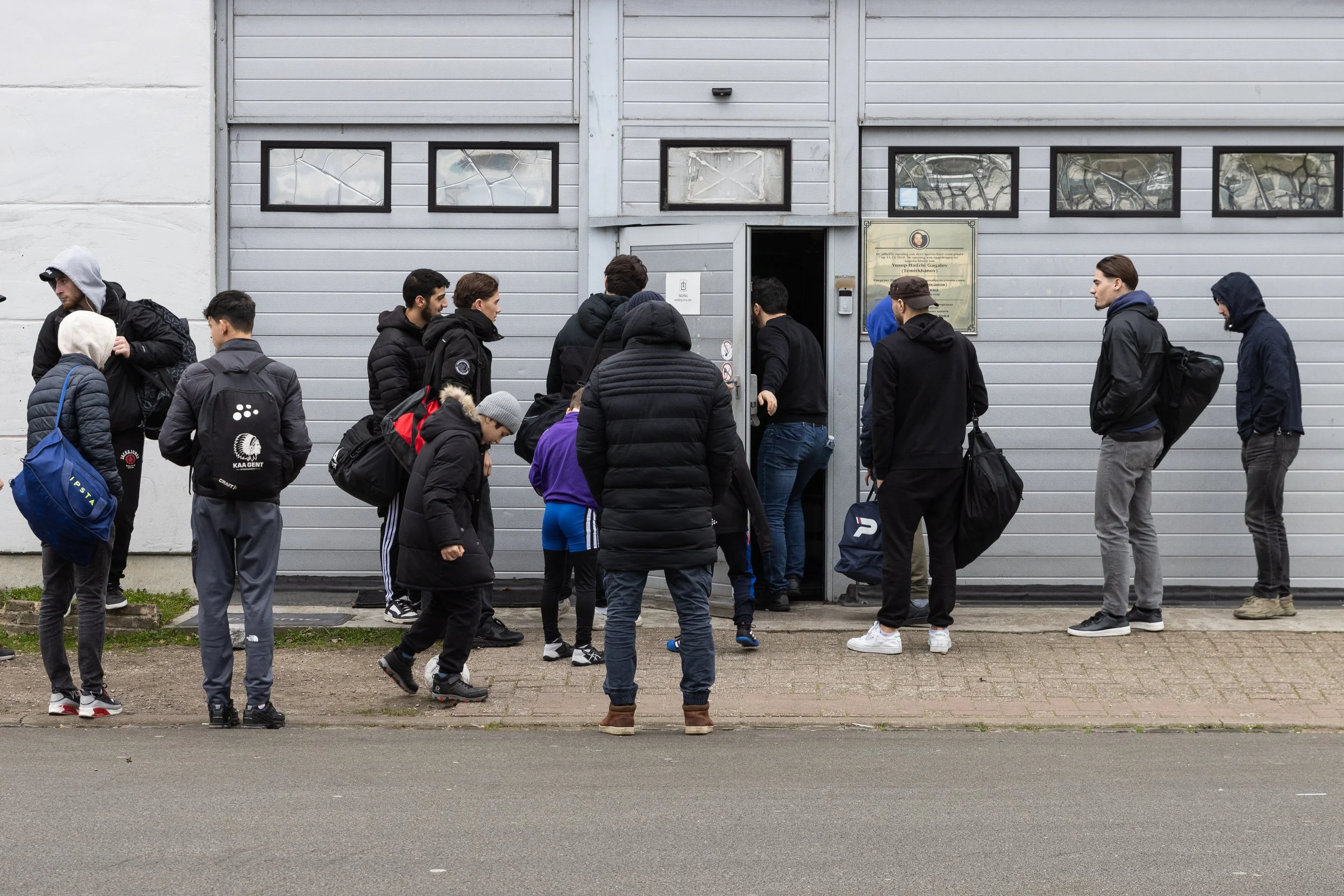 Een groep mensen staat in de rij voor een gebouw met een grijze gevel en vier ramen. Ze dragen winterkleding, backpacks en lopen naar binnen of wachten.