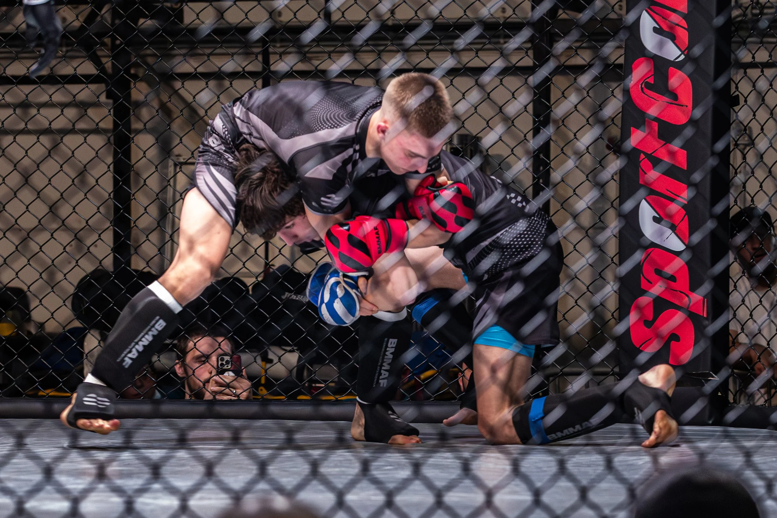 Twee MMA-vechters in de octagon, de ene bovenop de andere, tijdens een gevecht. Eén vechter draagt zwarte en blauwe shorts, de ander zwart met blauwe accenten. Beiden dragen handschoenen en zijn op de grond, omgeven door een metalen hek.