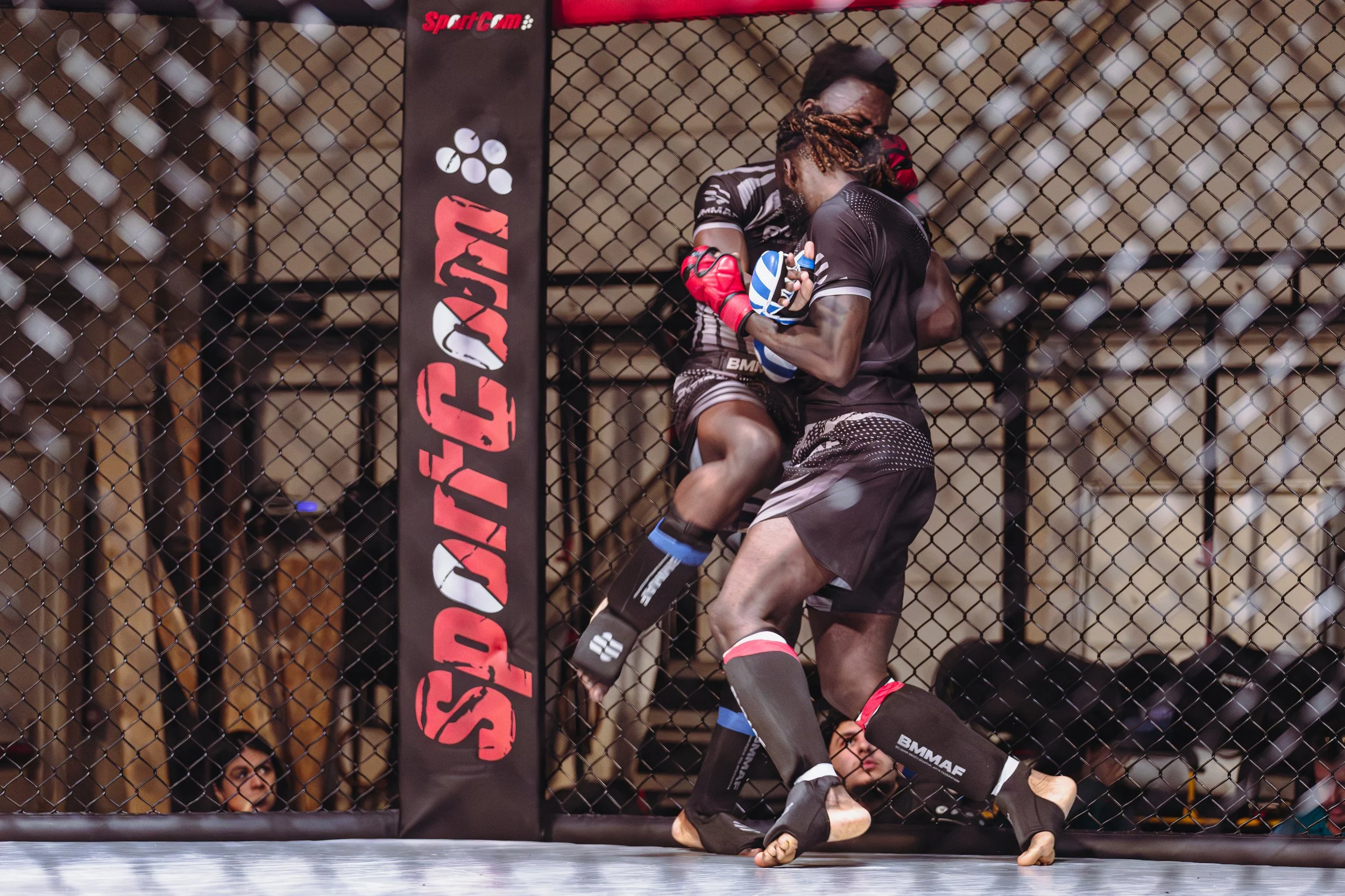 Twee MMA-vechters in een gevecht binnen een kooi, met een zwart hek en een mat met sponsors in de achtergrond.