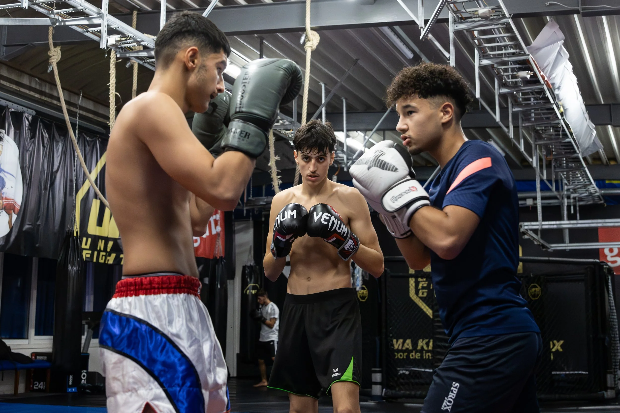 Drie jonge mannen staan in een bokstraining in een sportschool, klaar om te oefenen. De man links heeft handschoenen en een korte broek, terwijl de man rechts ook handschoenen draagt en een trainingsoutfit. De persoon in het midden kijkt geconcentreerd toe.