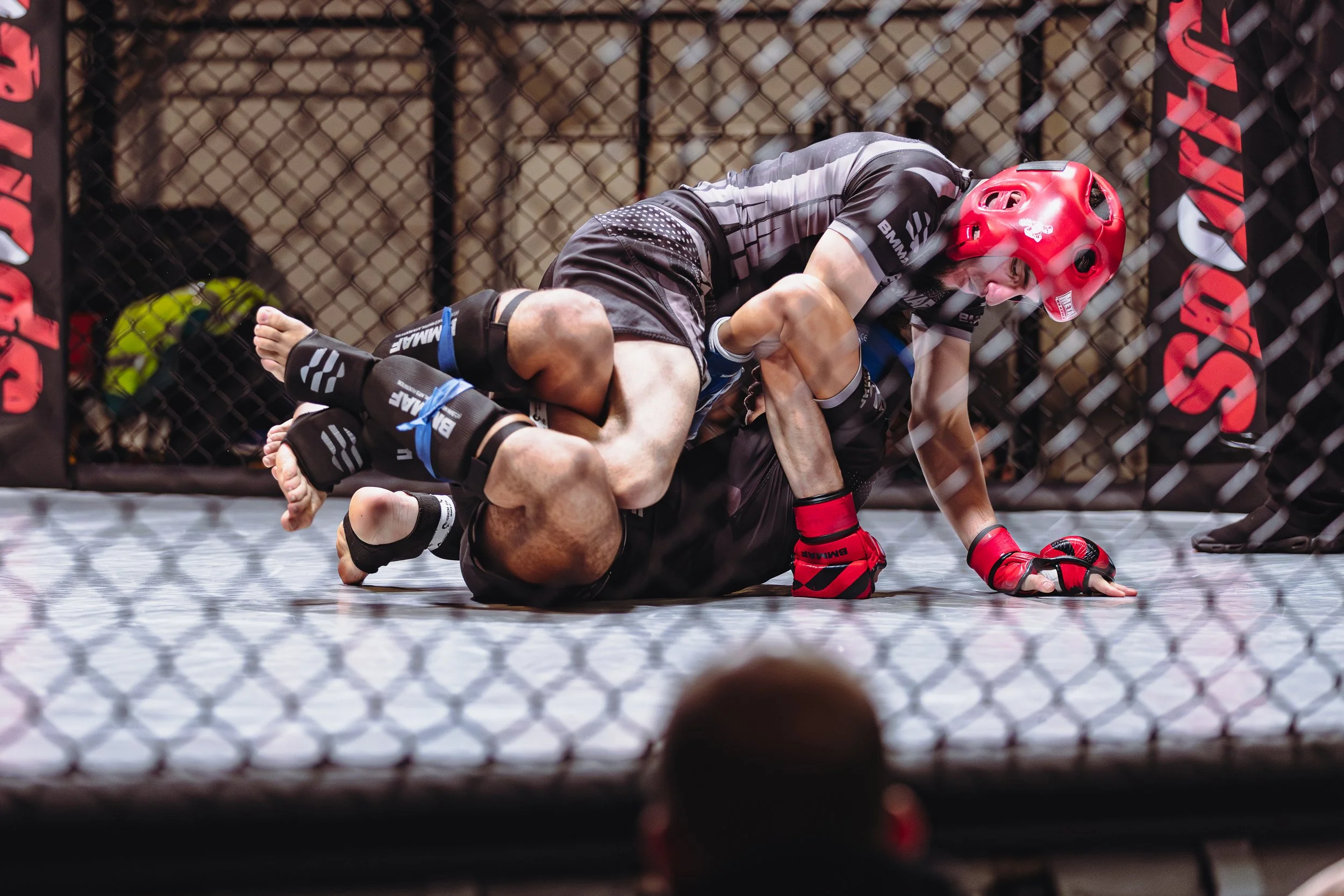 Twee MMA-vechters tijdens een gevecht in de octagon, met één die buitengewoon op zijn rug ligt en de ander die bovenop hem ligt, de winnaar draagt een rood hoofddeksel.