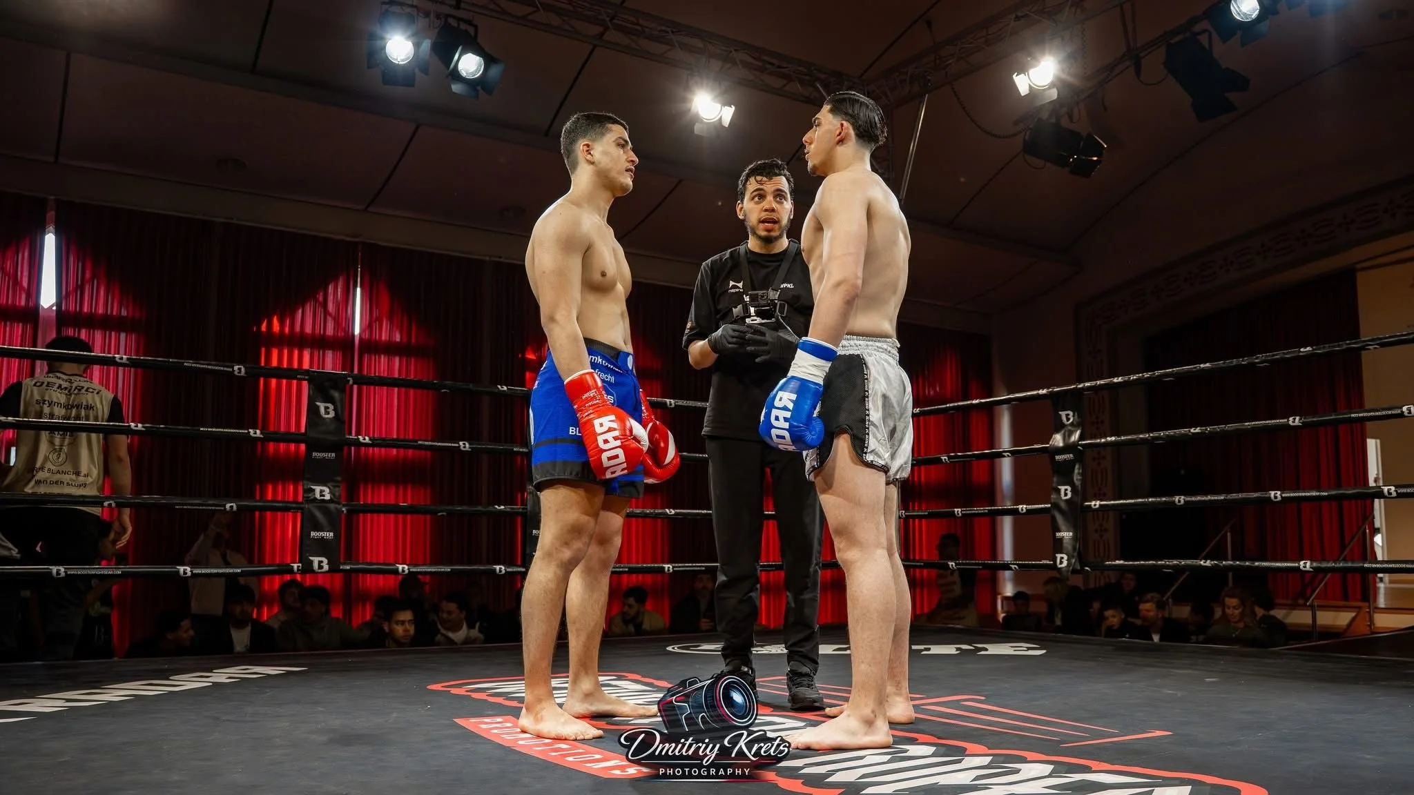 Twee boksers staan in de ring tegenover elkaar, klaar voor de wedstrijd, terwijl de scheidsrechter hen adviseert. Eén boksers draagt rood, de ander blauw, en er is publiek op de achtergrond.