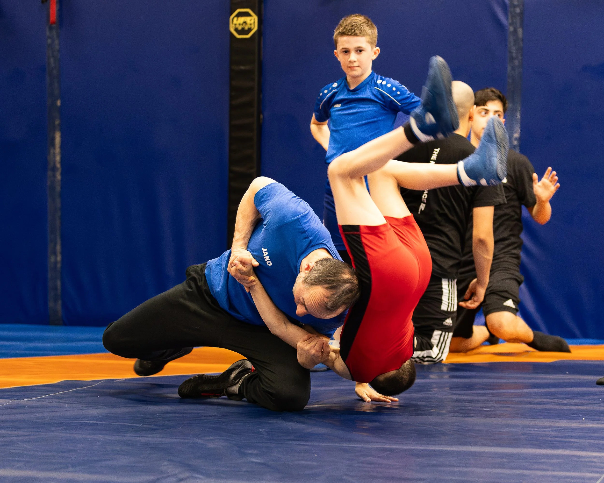 Twee jongens oefenen worsteltechnieken op een mat in een sportschool, één jongen ligt op de grond terwijl de andere hem vasthoudt.