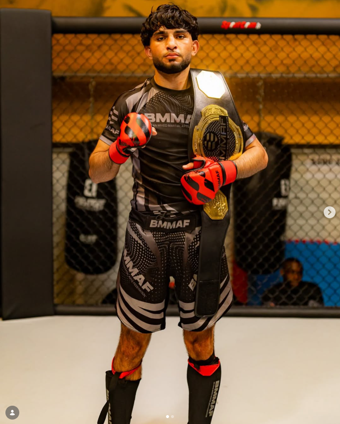 Een man in MMA-kleding en handschoenen, met een kampioensgordel om zijn schouder, staat binnen een MMA-kooi.