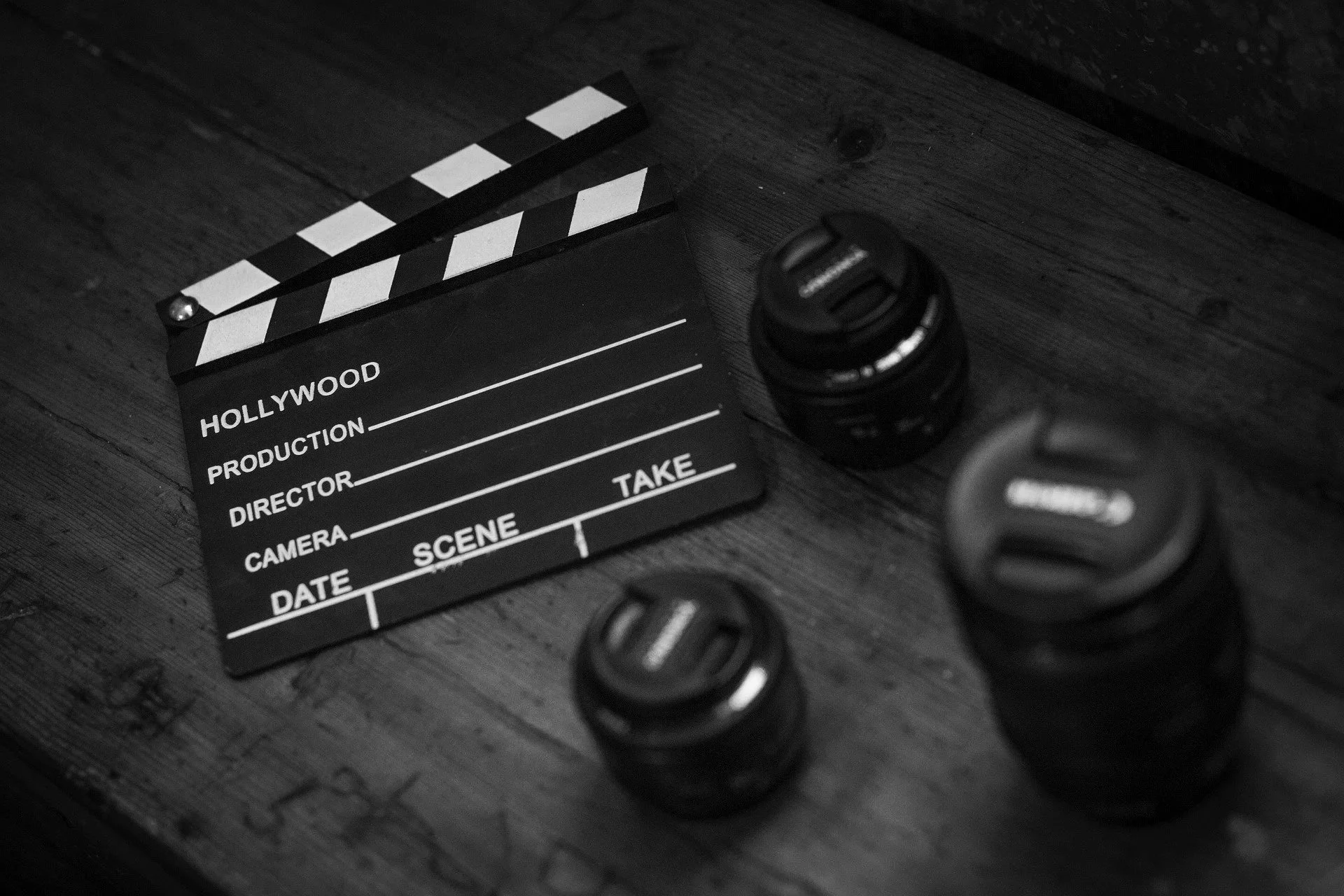 Black and white film slate with sections labeled Hollywood, Production, Director, Camera, Date, Scene, Take, and corresponding lines, along with three Canon camera lenses on a wooden surface.