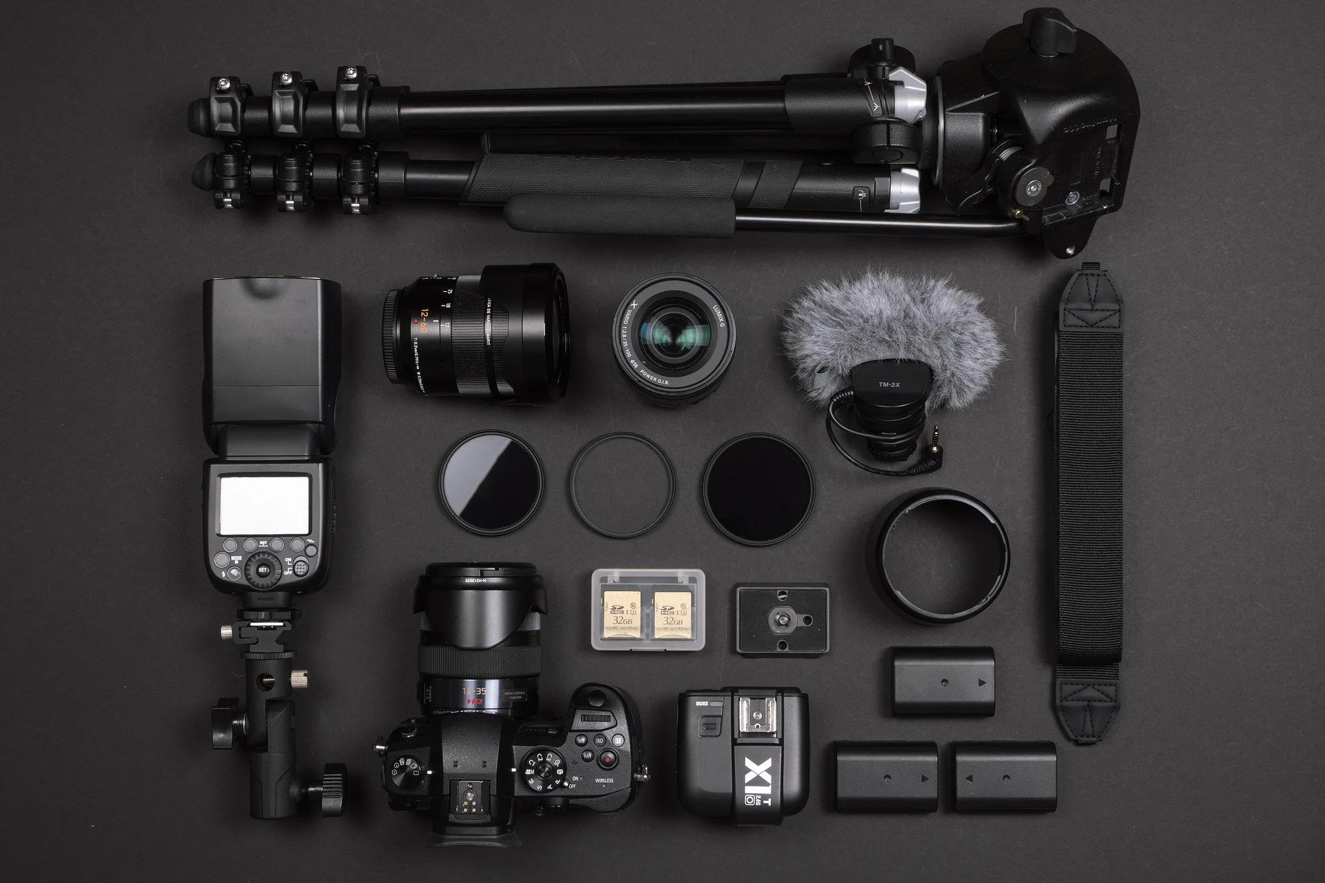 Photography equipment including a tripod, camera lens, camera body, flash, filters, SD cards, a camera with a mounted lens, a microphone, and other accessories on a dark background.