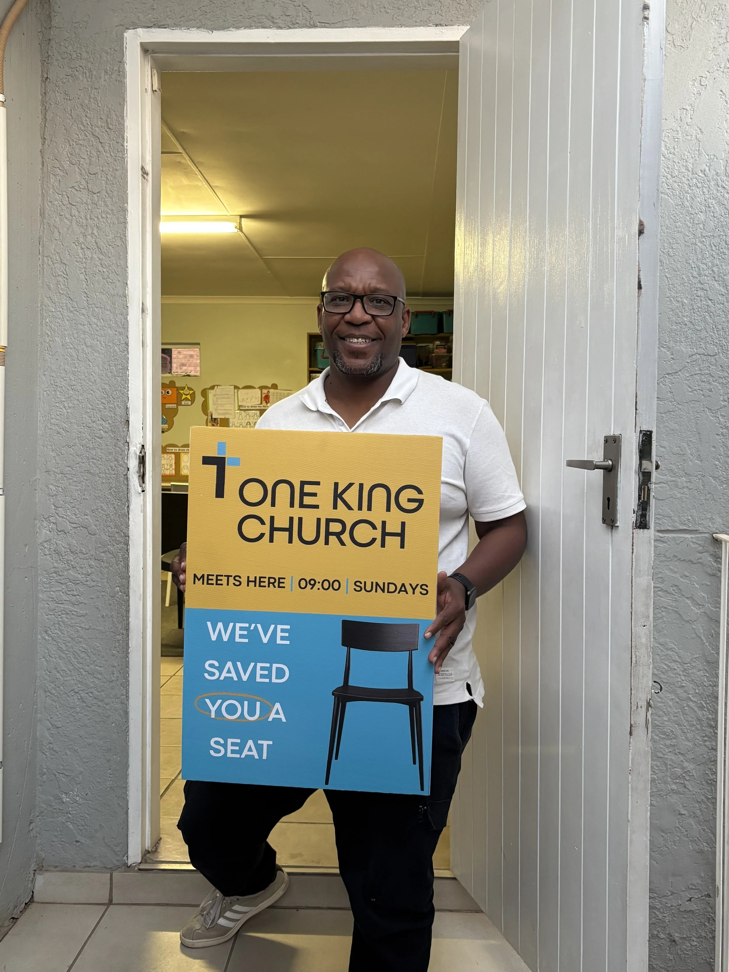 A man standing in a doorway holding a sign that reads 'One King Church meets here 09:00 Sundays We’ve saved you a seat', with a smile and glasses, inside a building with beige walls and a bulletin board in the background.