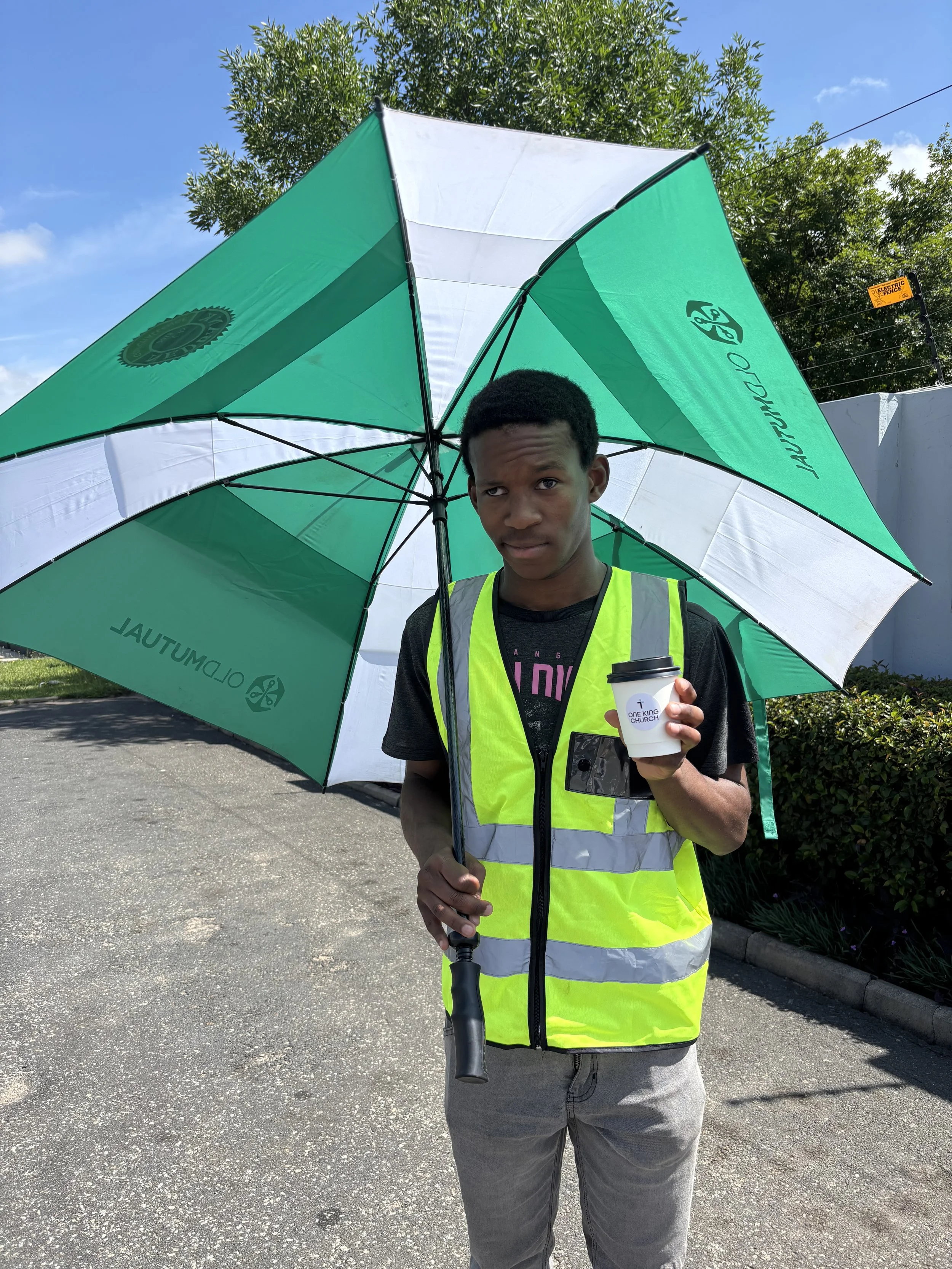 A young man wearing a high-visibility safety vest standing on a paved outdoor surface under a clear blue sky. He holds a large green and white umbrella with, and a small coffee cup with the logo 'One King Church.' Behind him are trees and a white wall with a yellow sign that reads 'Electric Fence.'