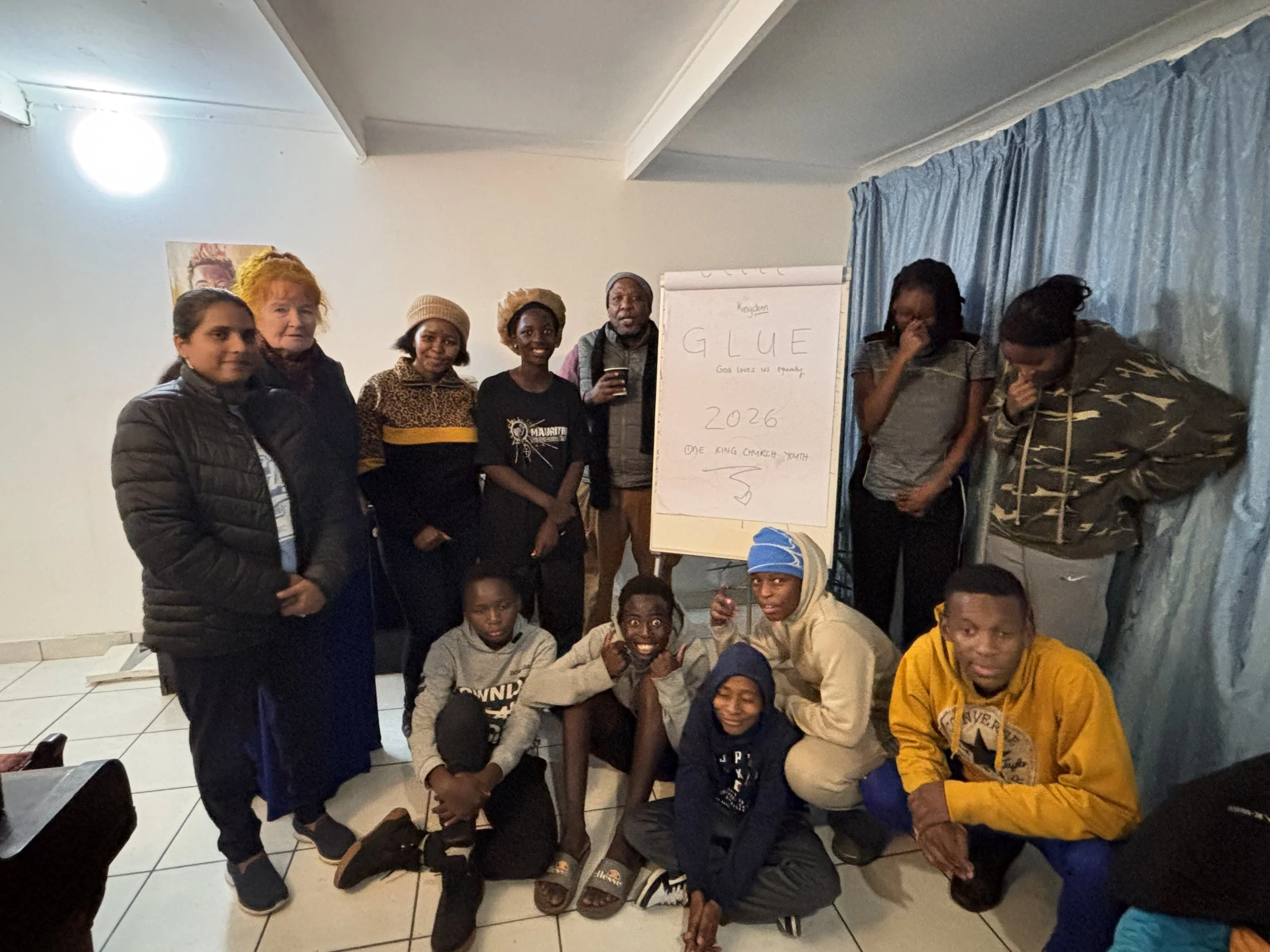 Group of people, both adults and children, gathered indoors around a whiteboard with the word "GLUE" (God Loves Us Equally) written on it, some smiling and others covering their faces or looking calm.