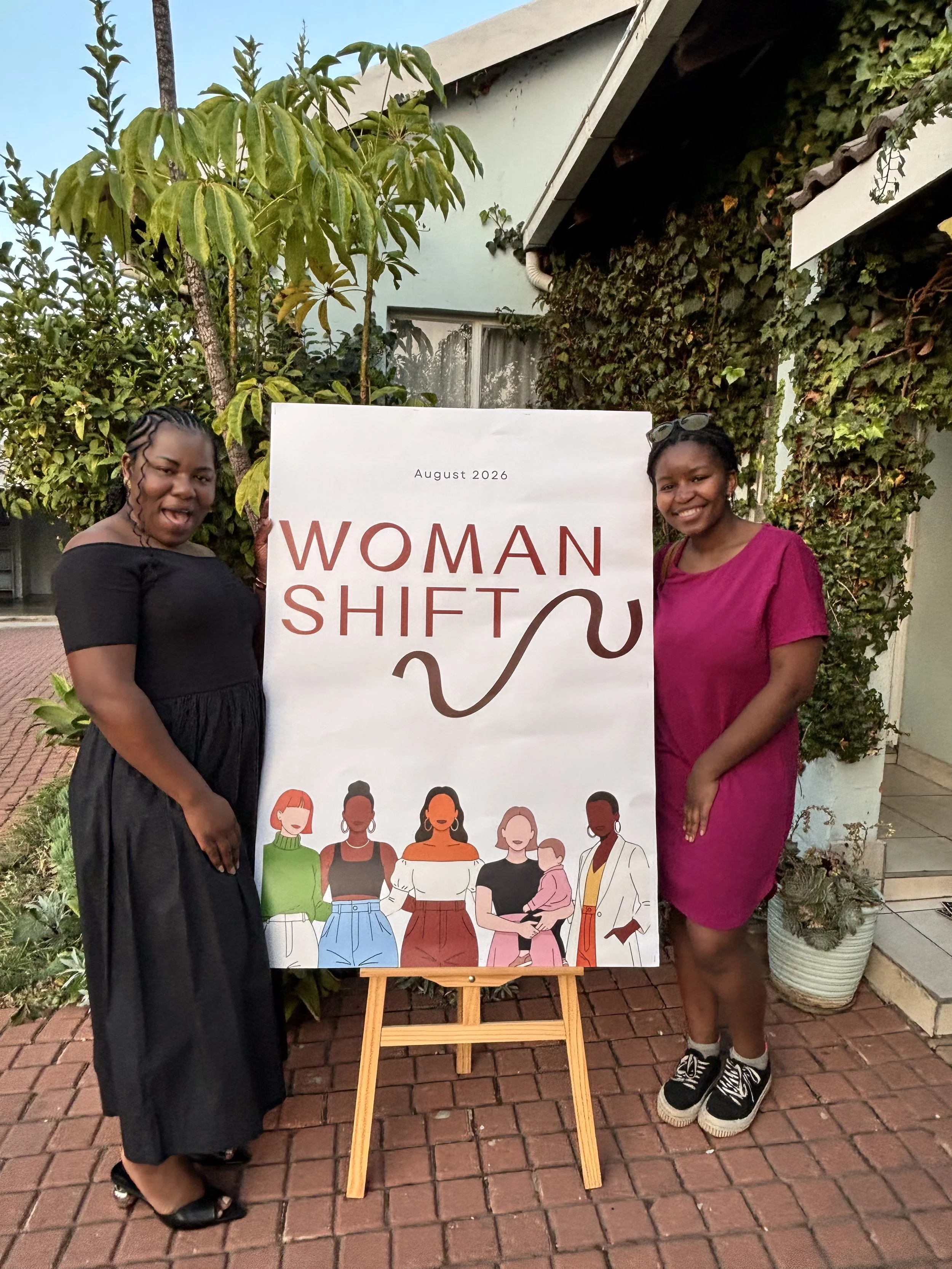 Two women standing on a brick pathway beside a sign that reads “Woman Shift”, one wearing a black dress with heels and the other a magenta dress with sneakers.