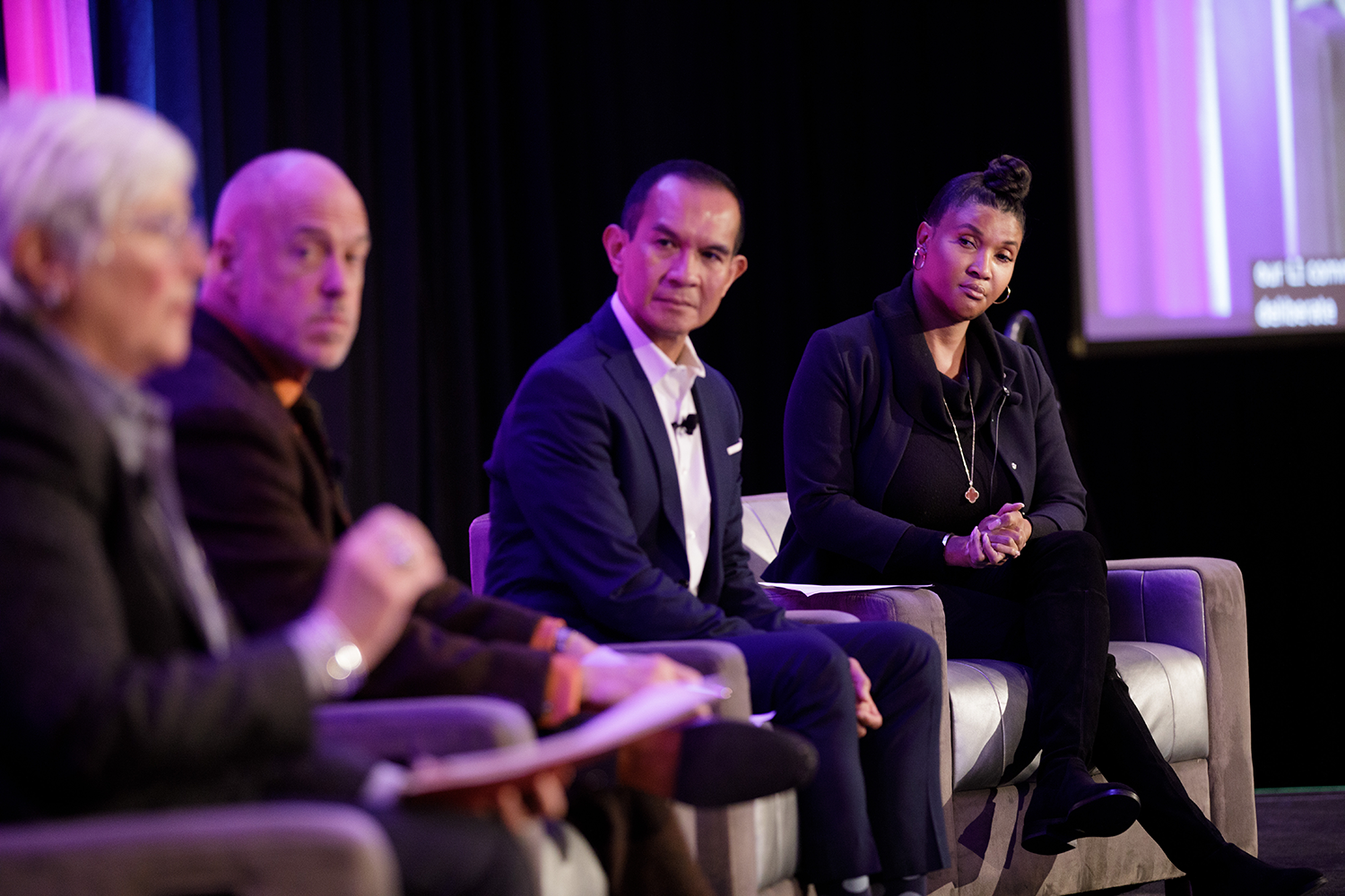 Panel of four people seated on stage during a discussion, with microphones clipped to their clothing, dark curtain backdrop, and a large screen on the right side.