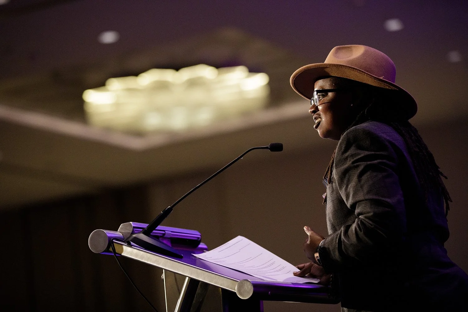 A person wearing glasses and a brown wide-brimmed hat speaking at a podium with notes, in a dark room with a blurred light fixture in the background.