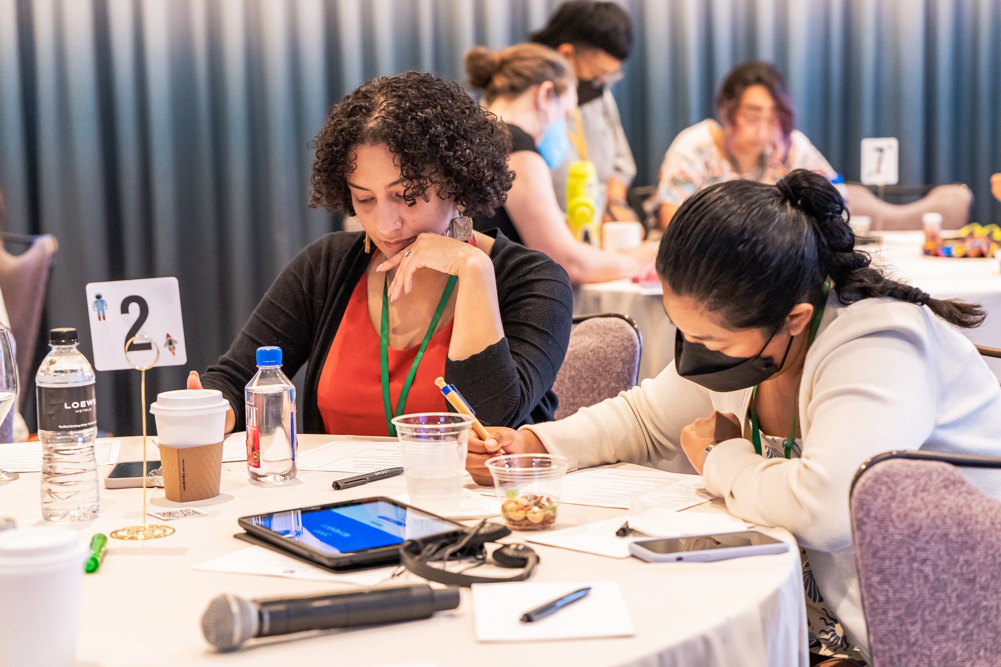 Two people sitting at a table, engaged in work or discussion, with notebooks, pens, and drinks, at a conference or workshop event.